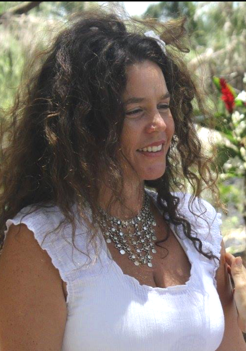 A woman with curly brown hair wearing a white top and layered silver jewelry, smiling outdoors in a garden setting with greenery and flowers.