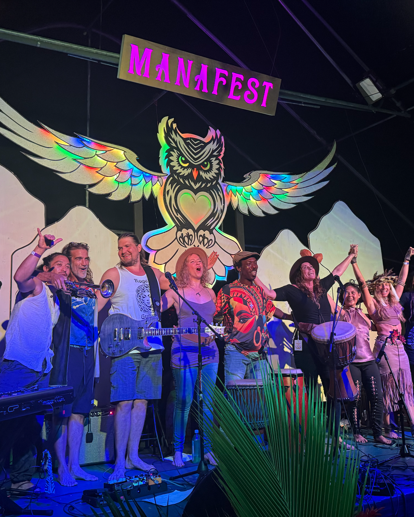A group of musicians and performers on stage at Mana Fest, with a colorful owl with wings as a backdrop and the Mana Fest banner overhead.