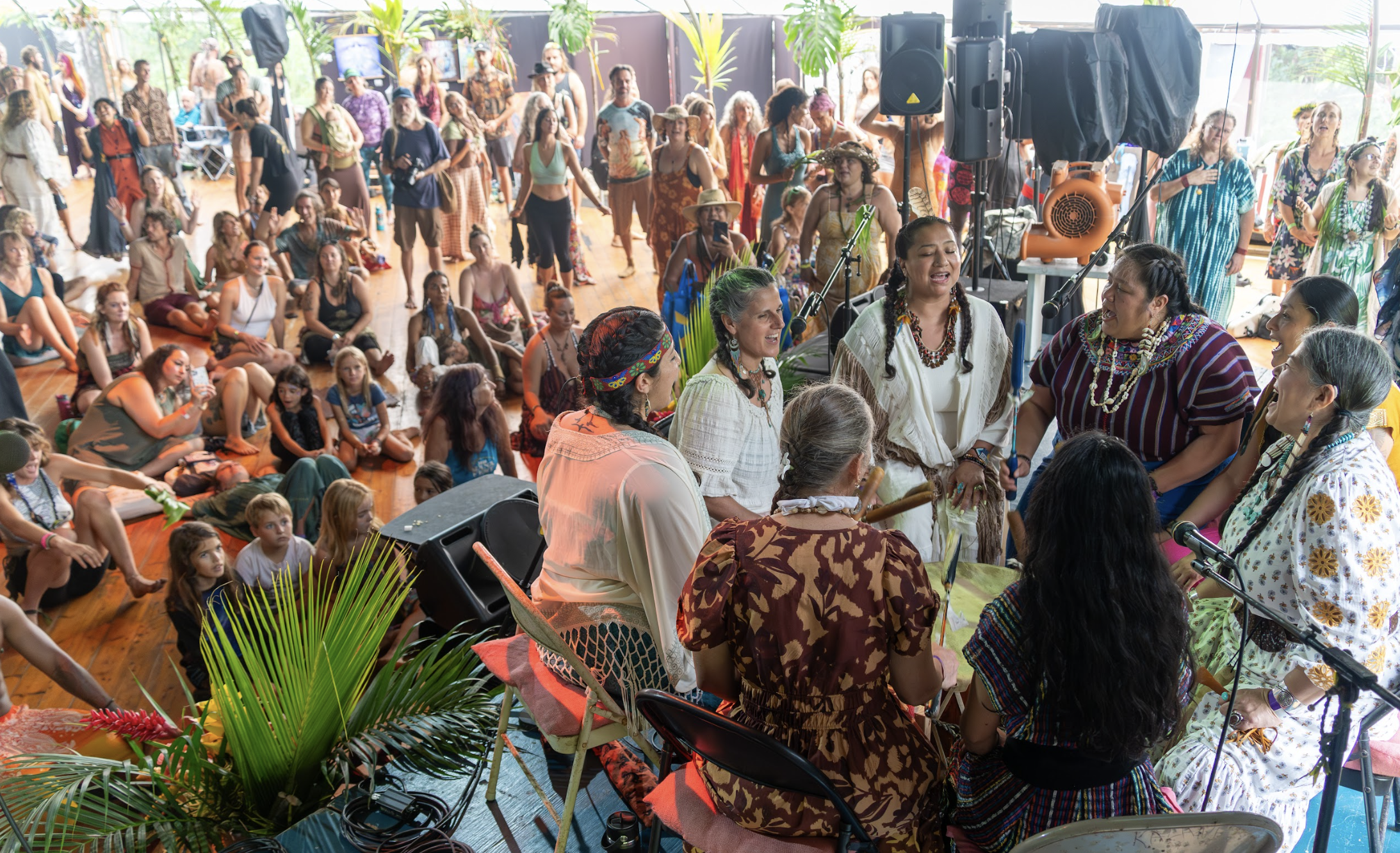 Group of women in traditional clothing performing music and singing in front of audience, with people dancing and sitting on the floor in a bright, tropical, indoor setting at  ManaFest, a transformational music, dance, arts, and culture festival