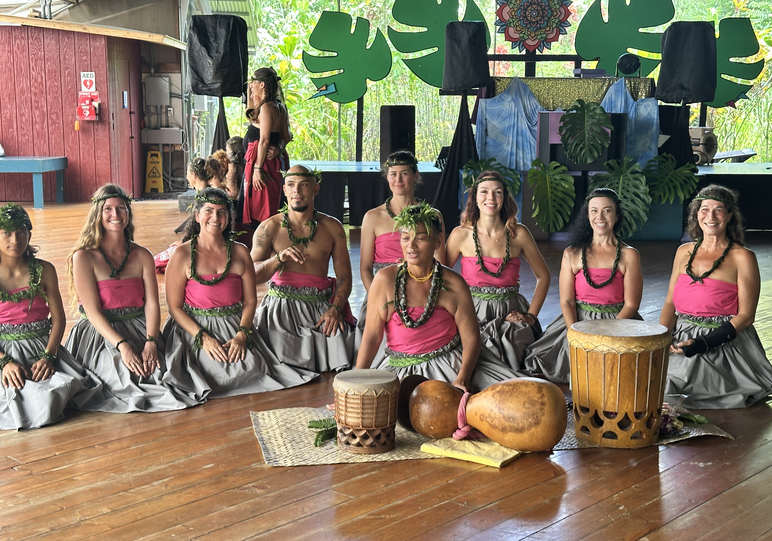 Group of Huka dancers in Hawiian cutural  attire with green ti leaf accessories, sitting on the floor with  Hawaiian musical drums.