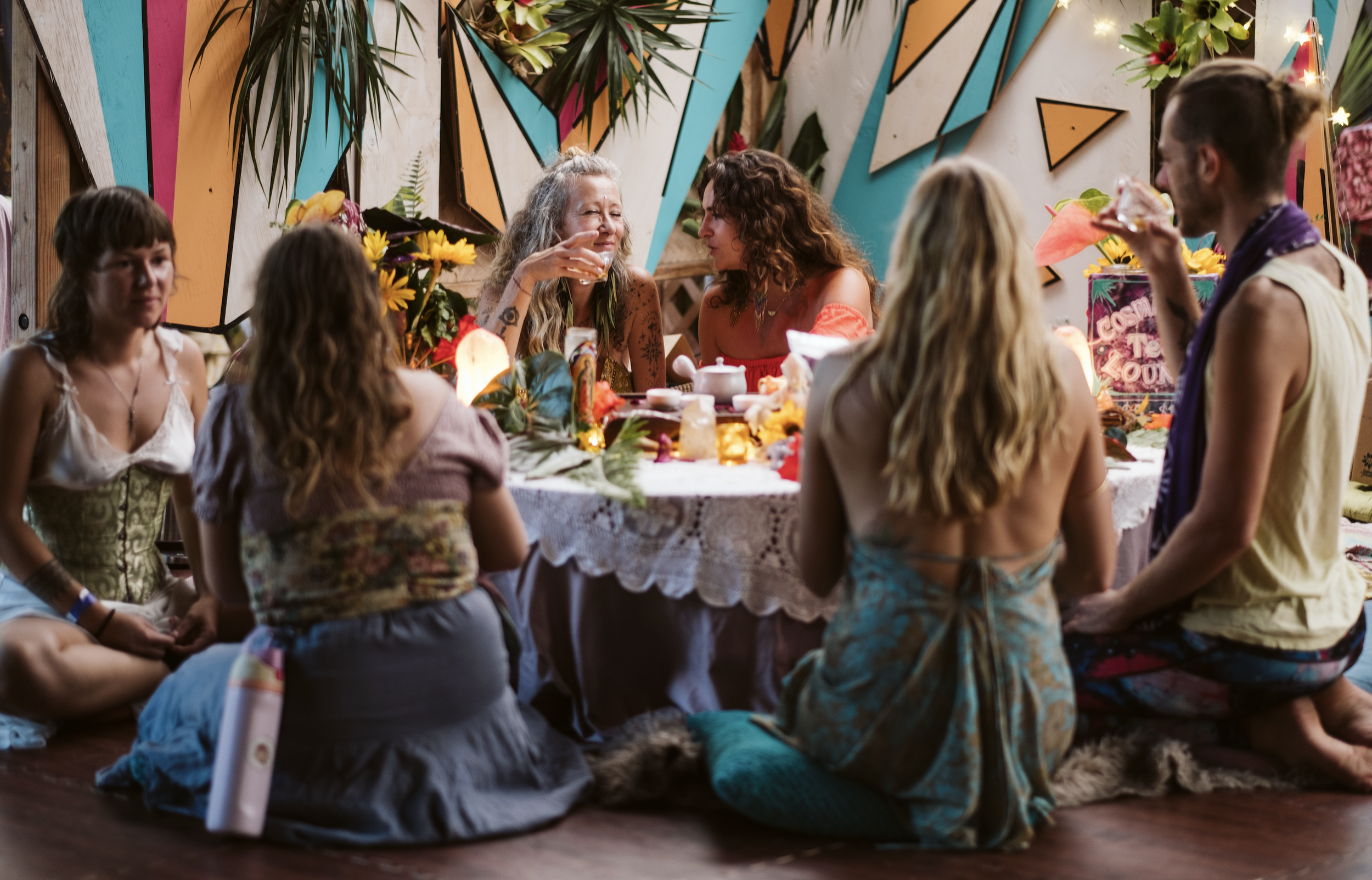 Group of people sitting around a decorated table at a party, with colorful geometric wall art and tropical plants in the background at  ManaFest, a transformational music, dance, arts, and culture festival on Big Island of Hawaii 
