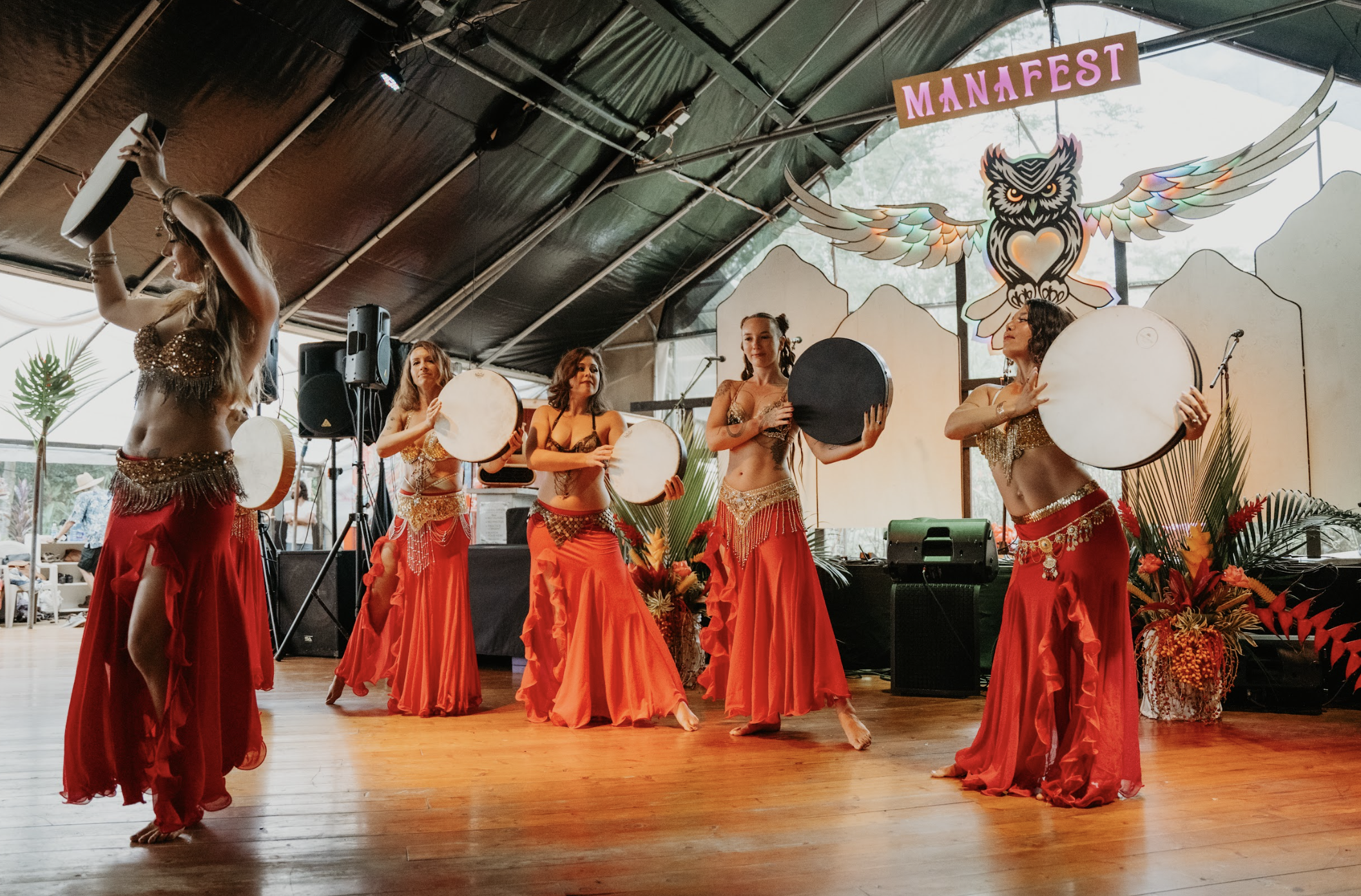 Six women in red and gold belly dance costumes perform with drums on a stage decorated with tropical plants, a colorful owl with wings, and a banner reading 'MANAFEST' at  ManaFest, a transformational music, dance, arts, and culture festival