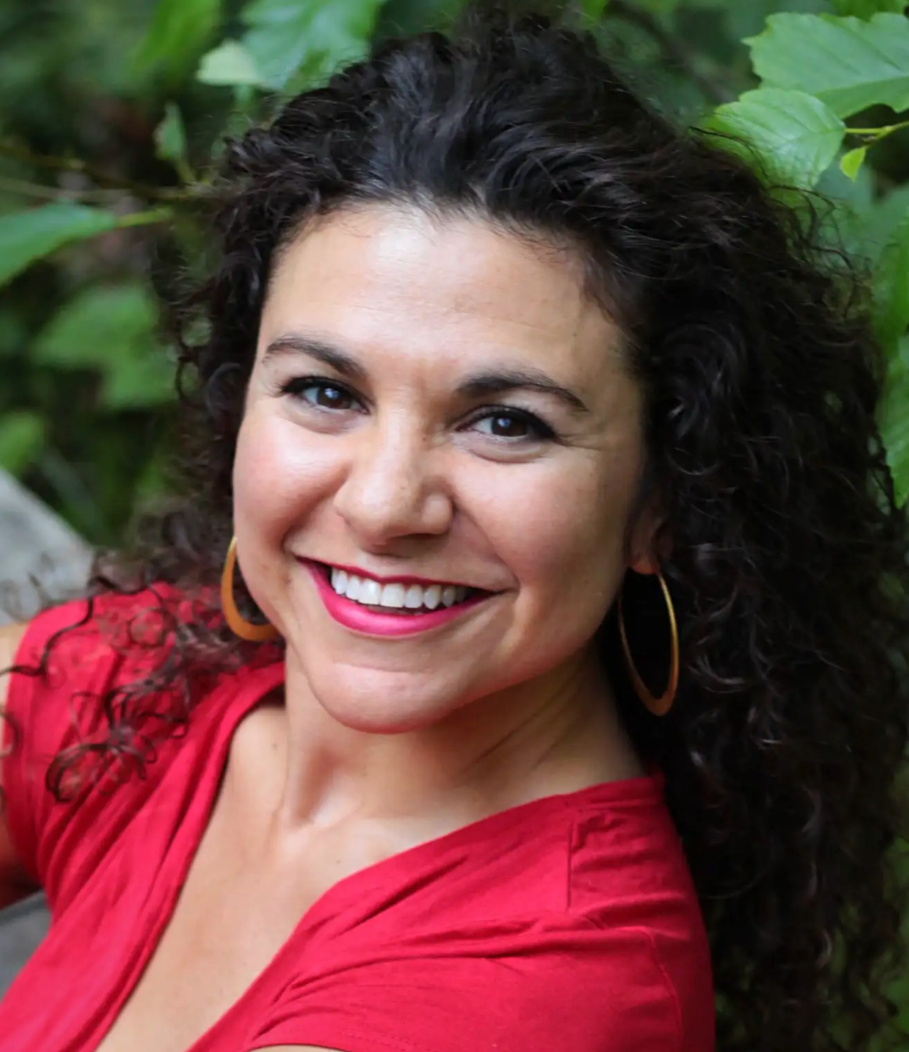 A woman with curly dark hair, wearing a red top and large gold hoop earrings, smiling outdoors with green foliage in the background.