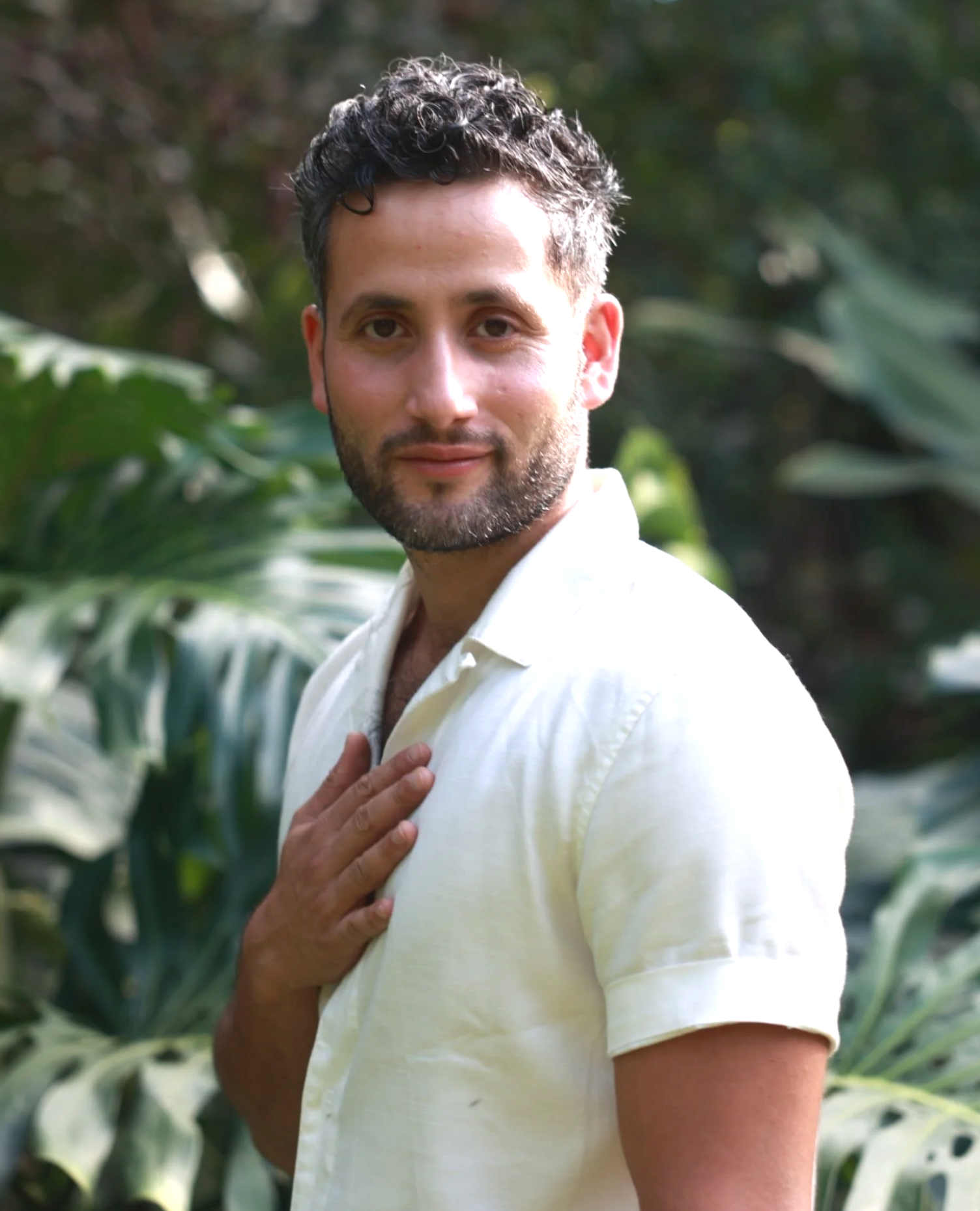 A man with dark, curly hair and a beard, wearing a light yellow shirt, is standing outdoors among large green plants, looking directly at the camera with a slight smile, his hand placed on his chest.