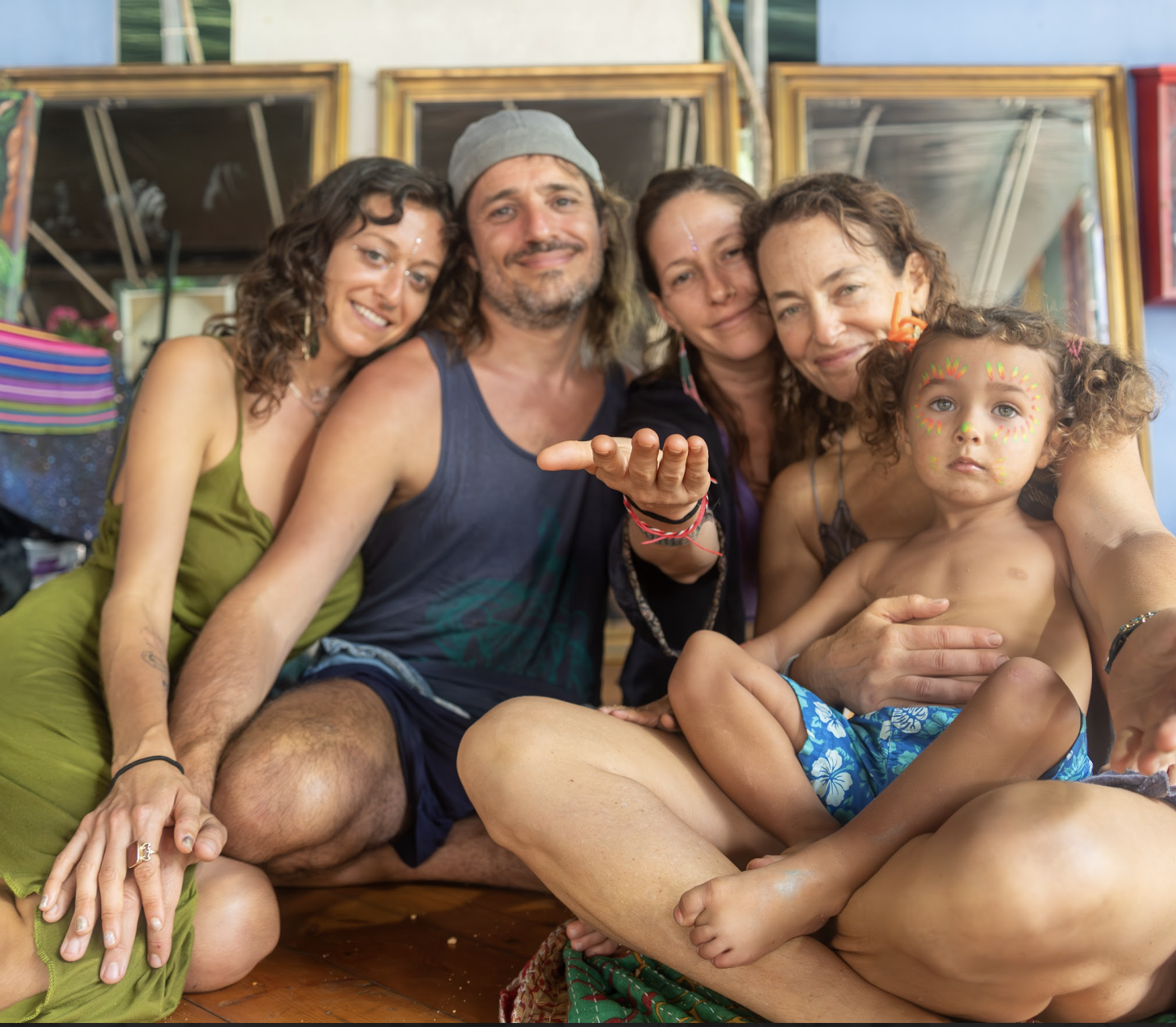 Group of diverse people, including a child with face paint, sitting close together indoors in front of mirrors at  ManaFest, a transformational music, dance, arts, and culture festival on Big Island of Hawaii 