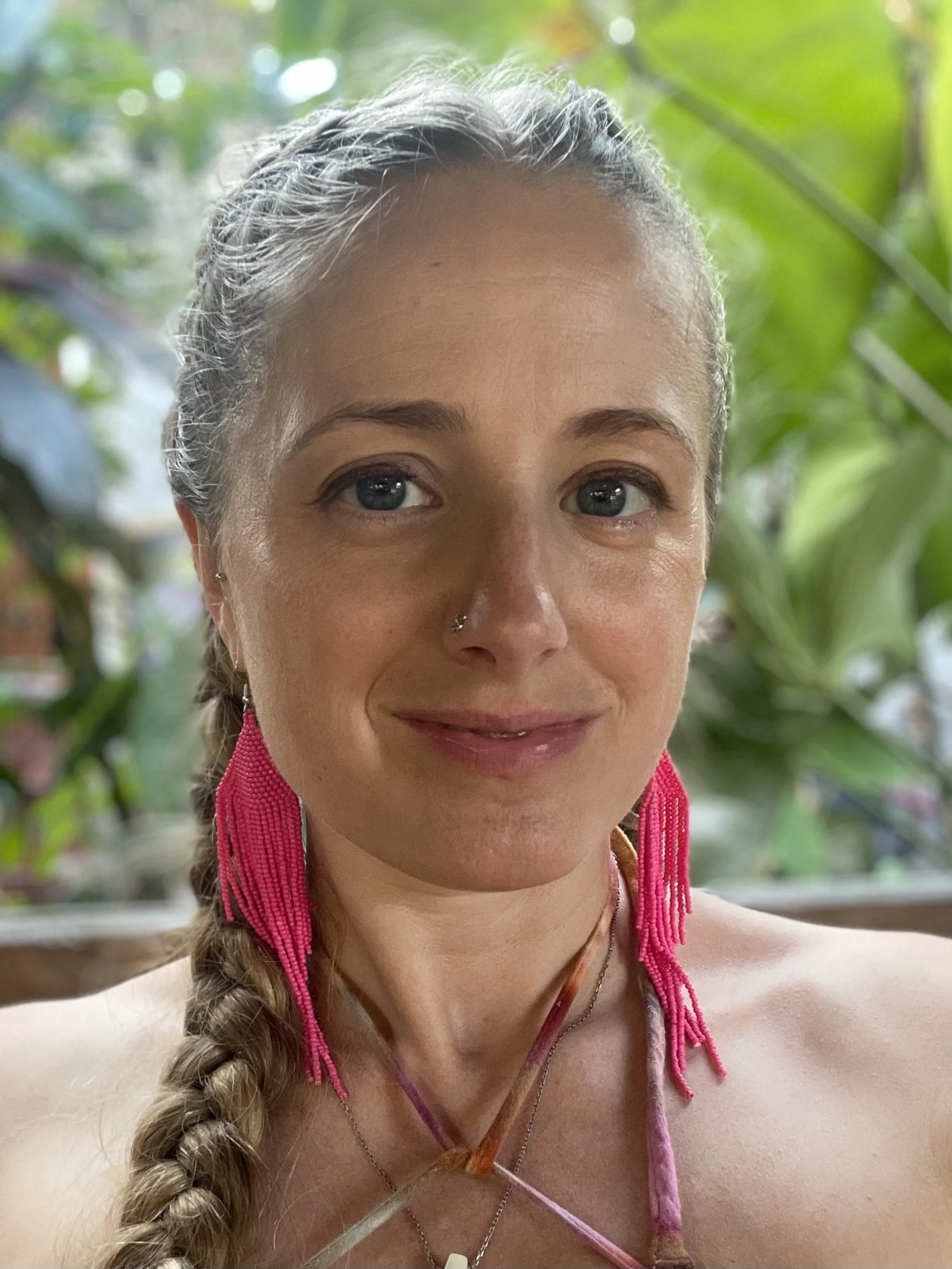 A woman with fair skin and braided blonde hair, wearing large pink beaded earrings and a nose ring, smiling and looking at the camera. She is outdoors surrounded by lush green plants.