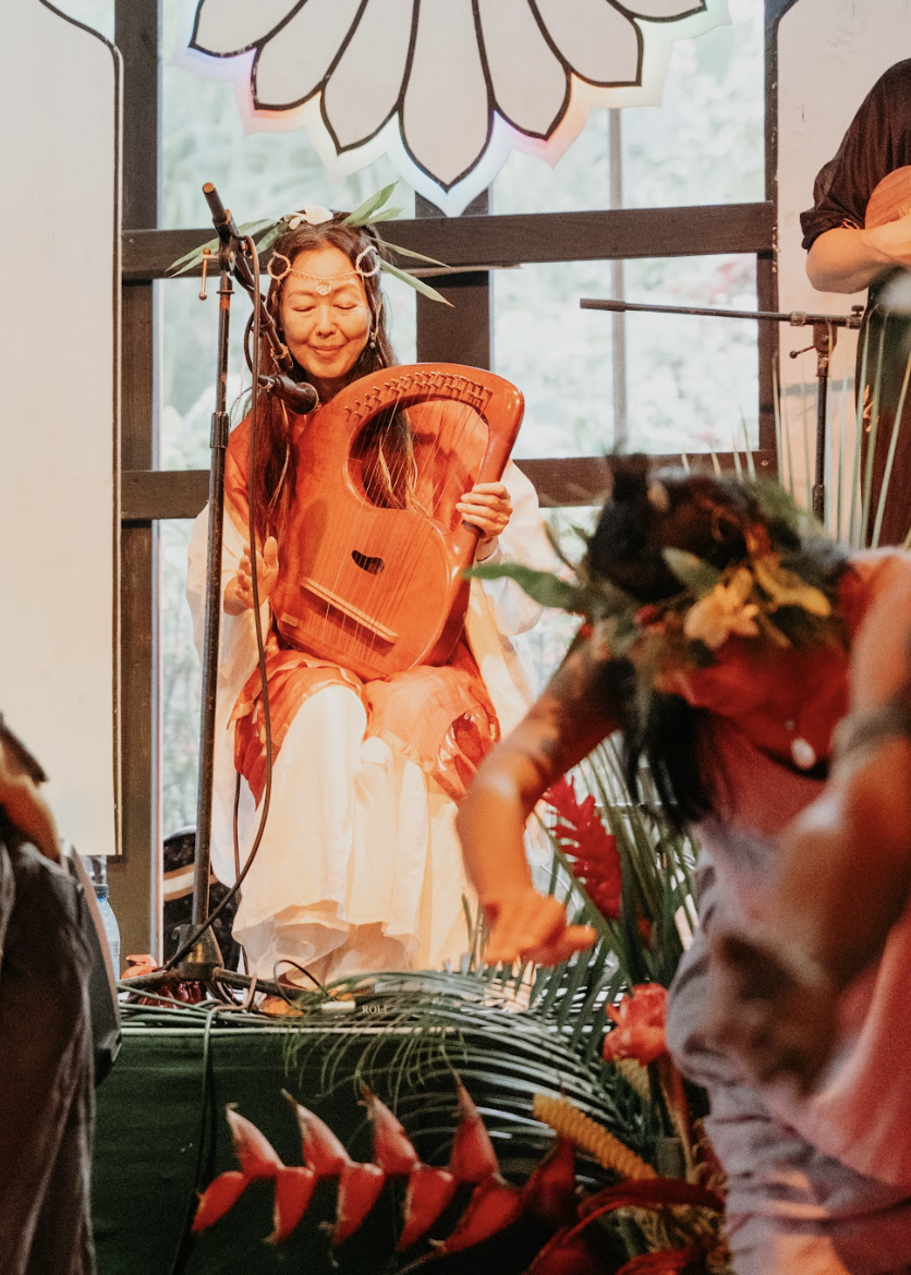 A woman dressed in traditional Hawaiian attire playing a wooden musical instrument on stage during a performance, at  ManaFest, a transformational music, dance, arts, and culture festival on Big Island of Hawaii 