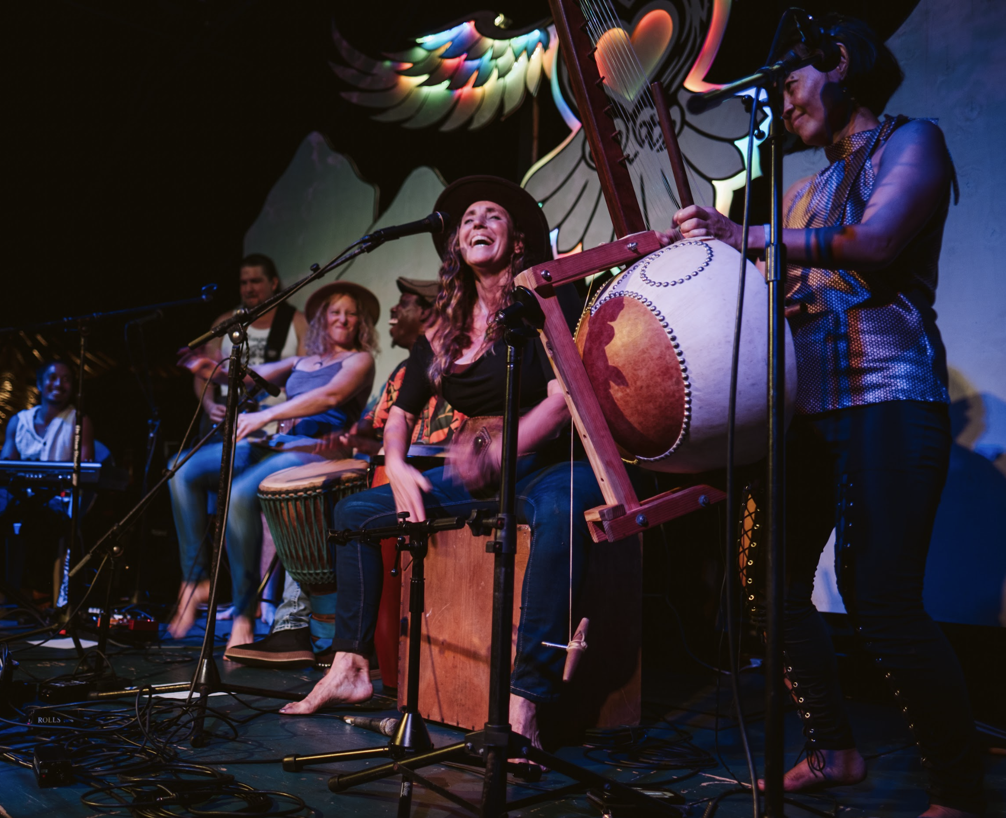 A group of musicians performing with Hope Medford on stage with colorful lighting and a winged heart mural in the background, including a woman playing a large drum and singing, and others playing percussion instruments at  ManaFest