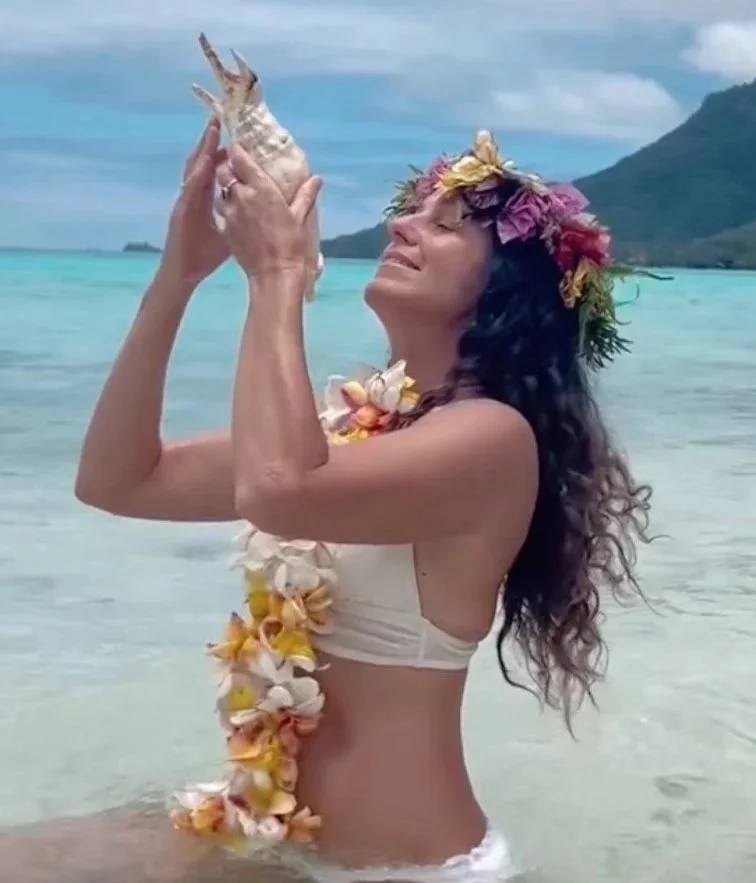 Woman in a bikini with a colorful flower lei and a flower crown, holding a seashell up in the ocean with mountains in the background.