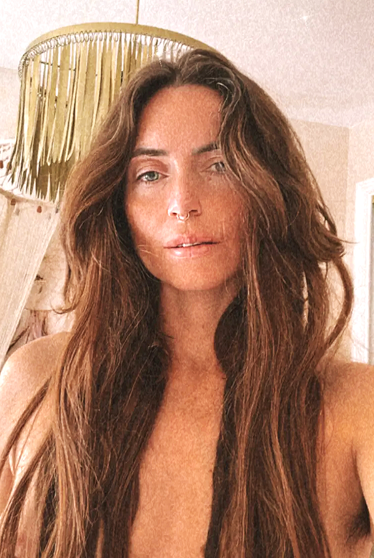 A woman with long wavy brown hair, fair skin, and blue eyes taking a selfie indoors. She has a septum piercing and is near a wooden and wicker lamp with a green fringe, in a room with beige walls.