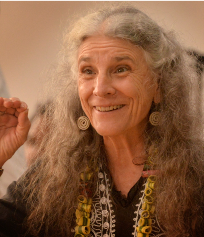A smiling elderly woman with long, curly gray hair, wearing spiral earrings and a black embroidered top, appears to be in a cheerful or excited mood.