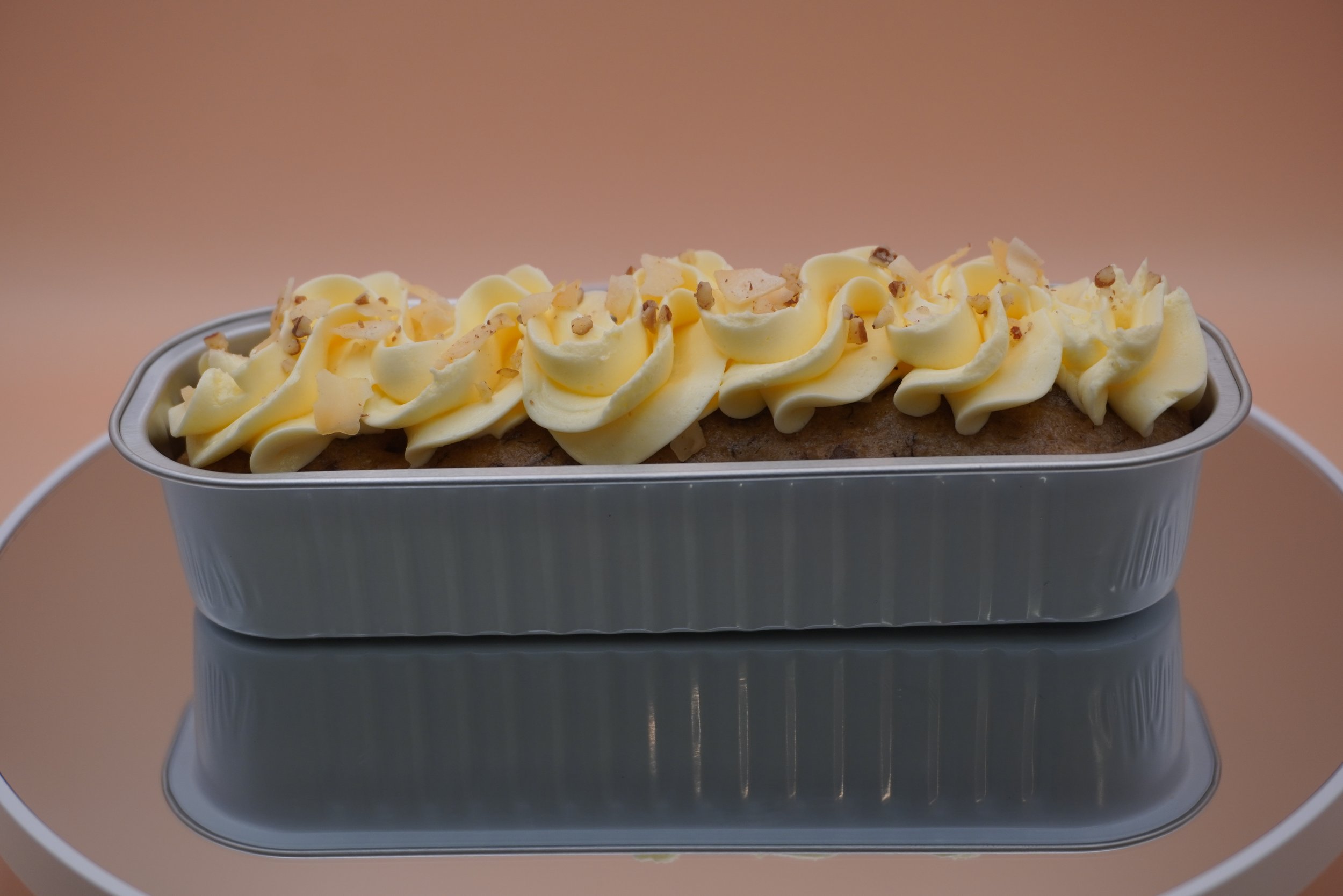 A loaf of bread topped with swirls of pale yellow frosting and sprinkled with chopped nuts, inside a rectangular metal baking pan.