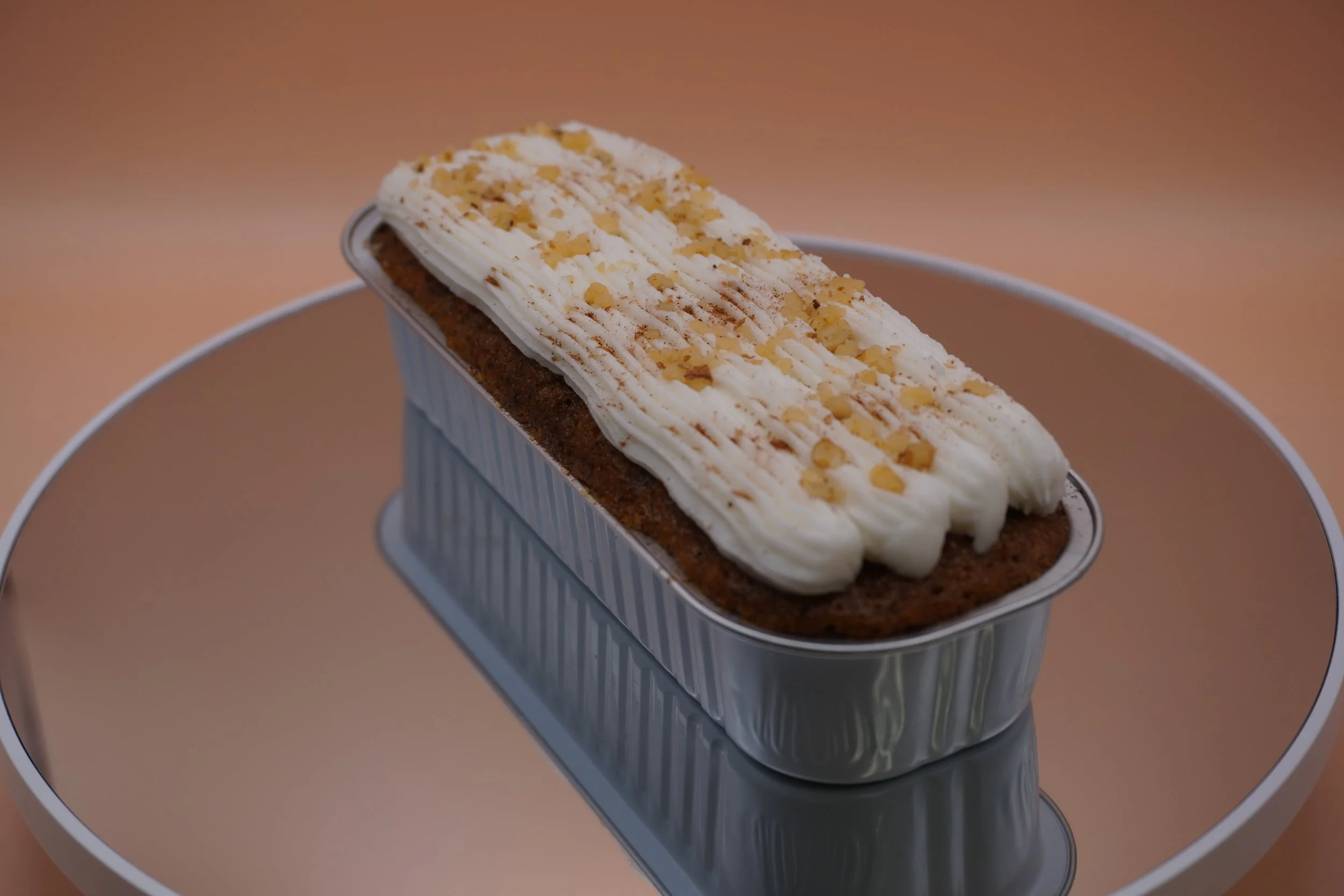 Chocolate loaf cake topped with icing and chopped nuts, in a metal pan, placed on a reflective surface.
