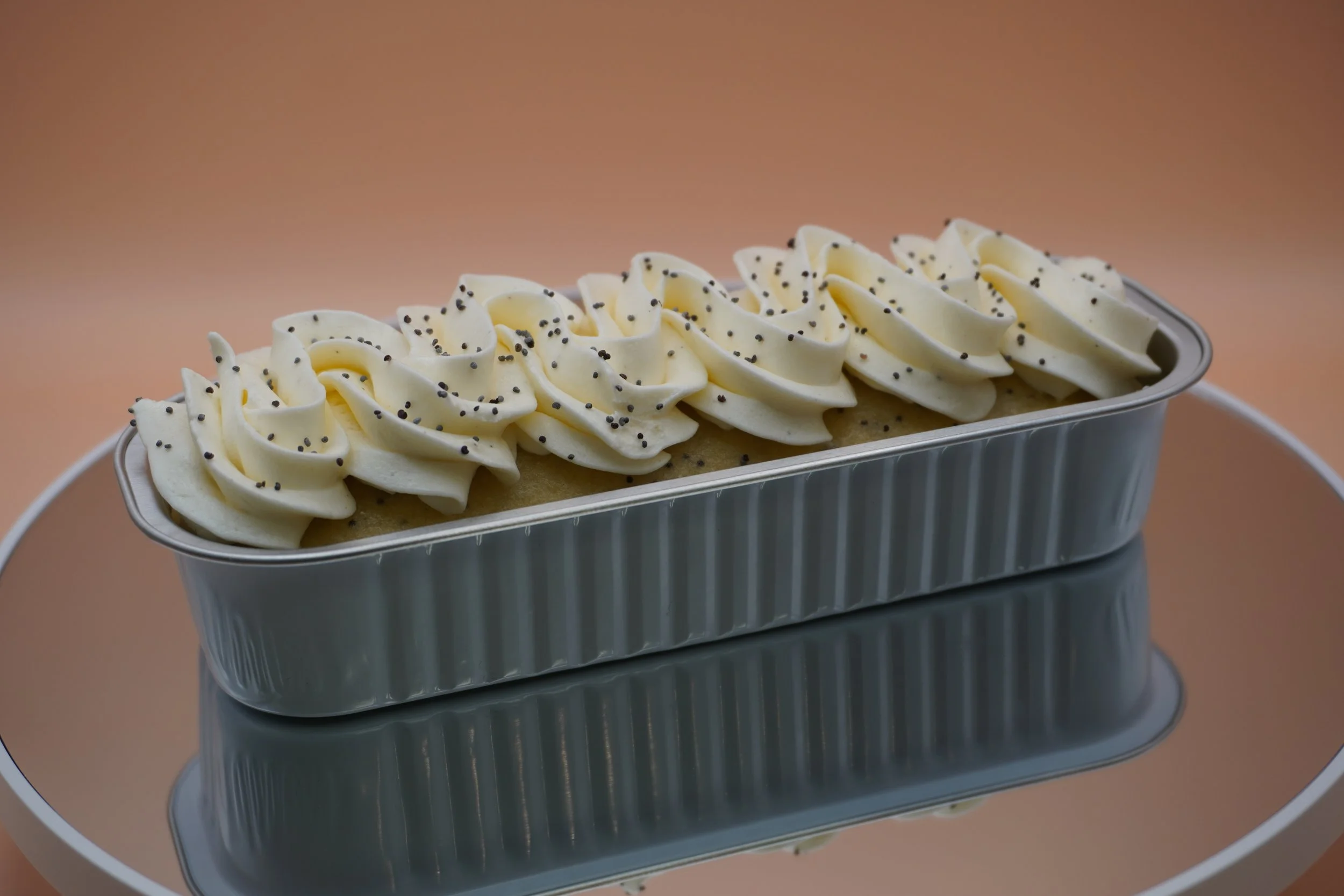 A loaf of baked cake topped with white frosting and black sprinkles in a metallic pan, placed on a reflective surface.
