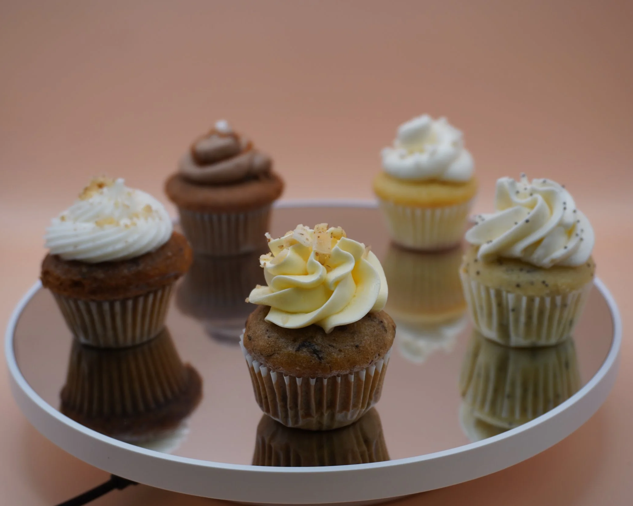 Six assorted cupcakes on a white reflective tray against a soft pink background.