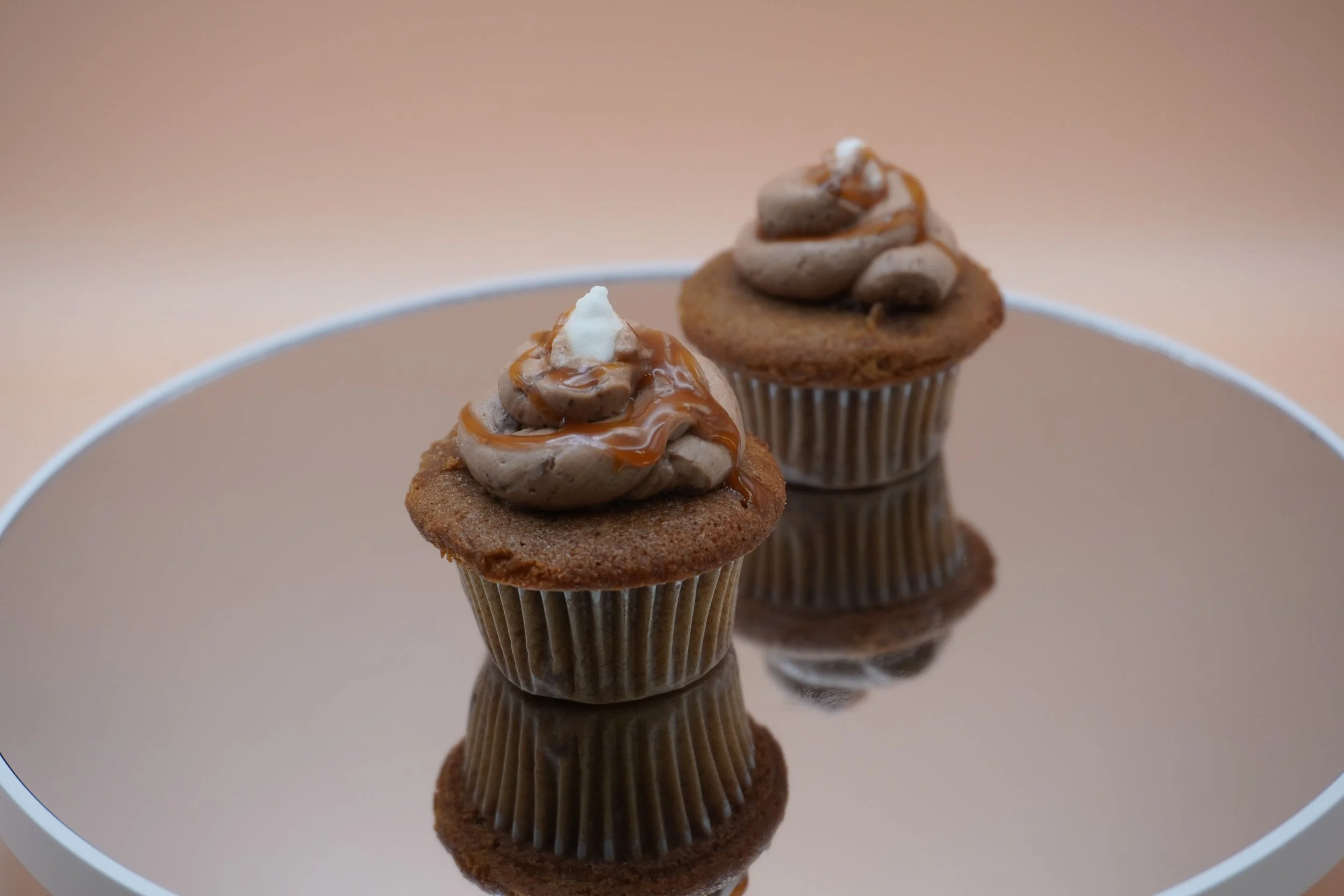 Two chocolate cupcakes with swirled frosting topped with caramel drizzle and small dollops of white cream, placed on a reflective surface.