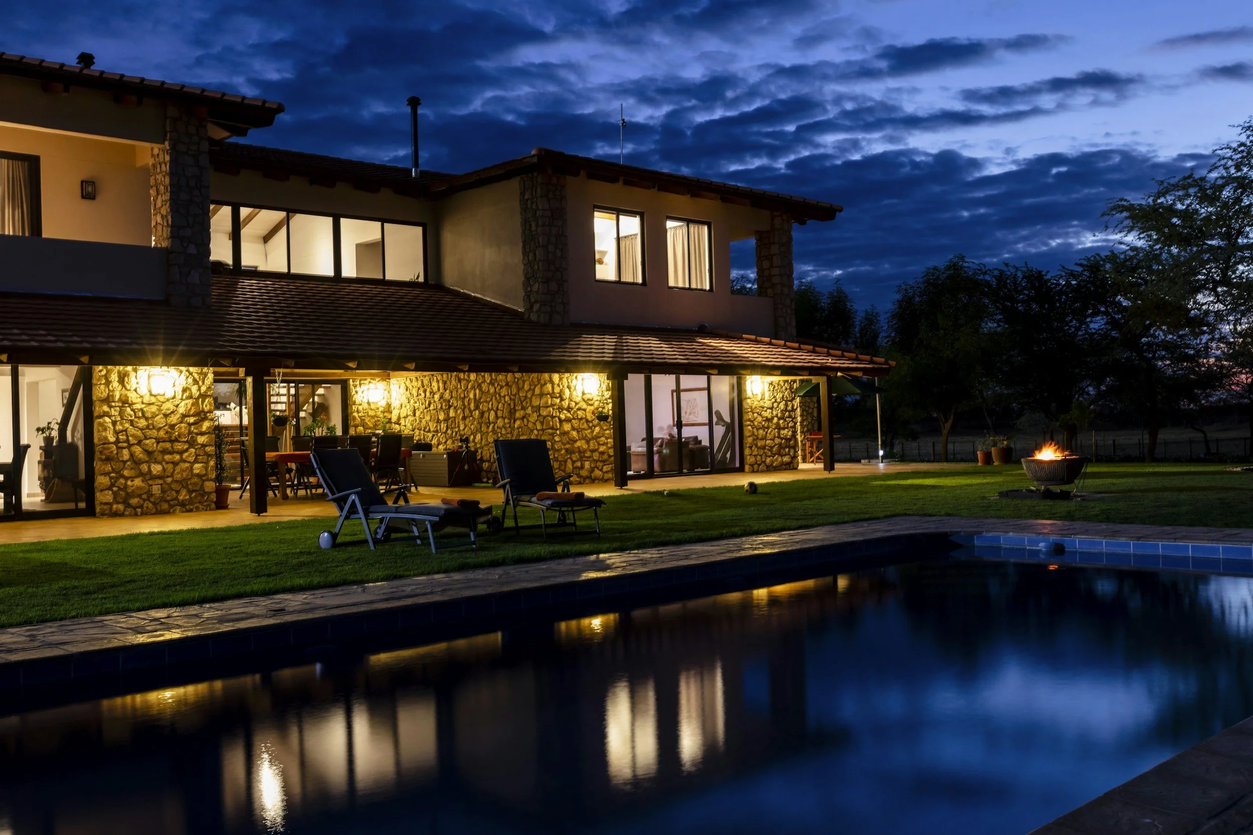 A two-story house with stone and stucco exterior, large windows, and a tiled roof, illuminated with outdoor lighting, with a backyard featuring patio furniture, a fire pit, and a swimming pool, under a cloudy evening sky.