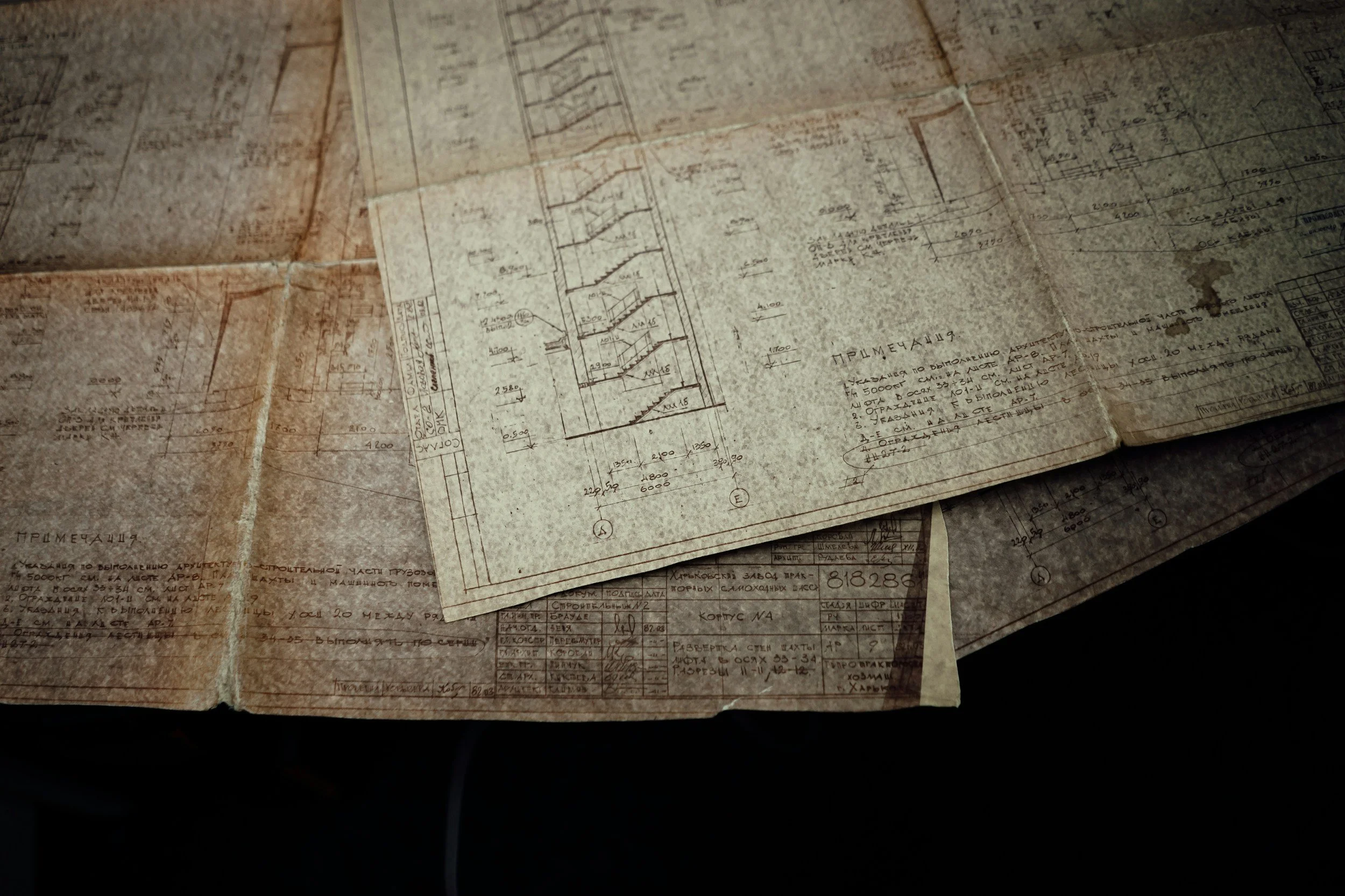 Old, worn architectural blueprints and technical drawings spread out on a surface, featuring detailed sketches, measurements, and handwritten notes.