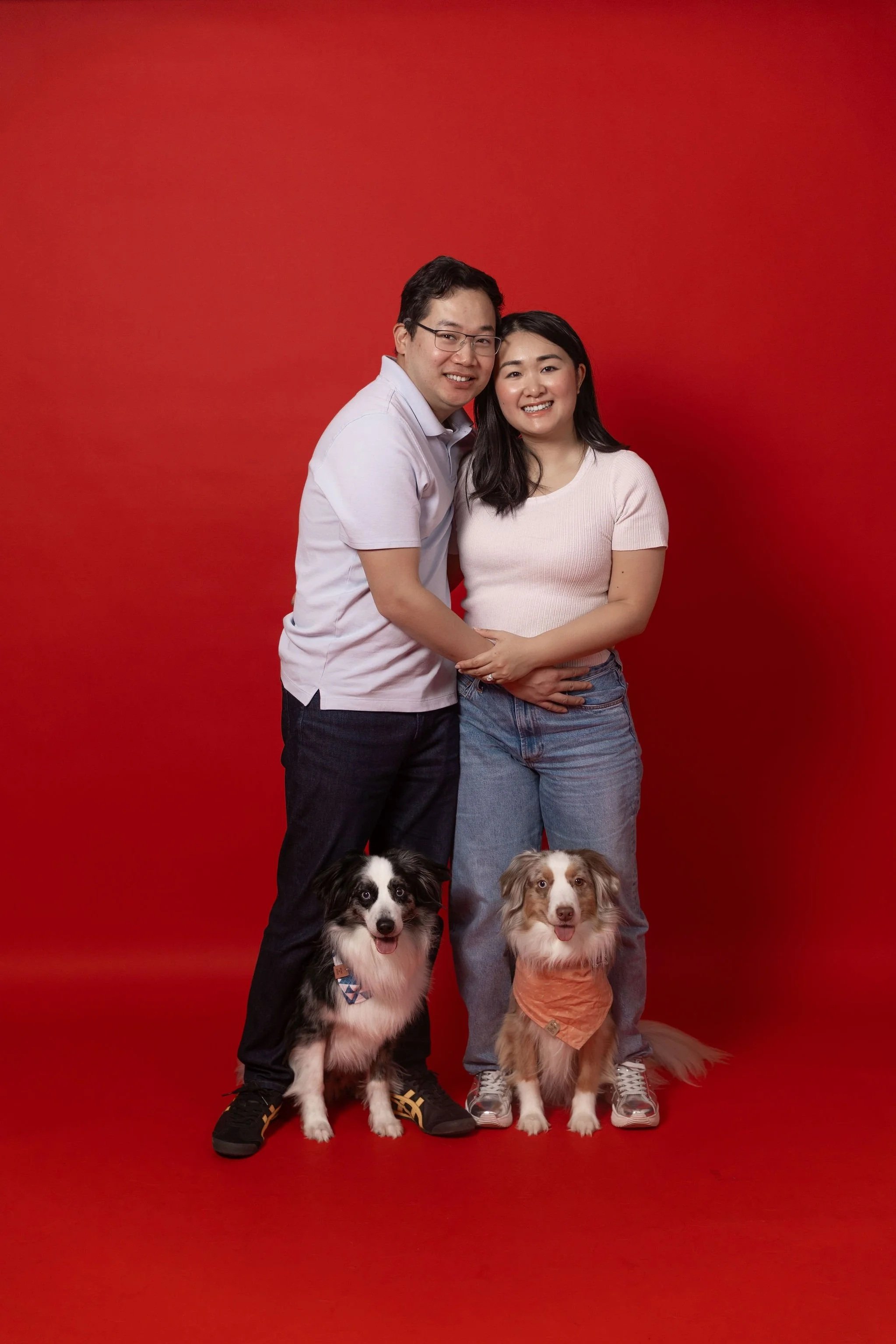 A smiling couple stands closely together against a red background, with a black and white dog and a brown and white dog sitting in front of them.