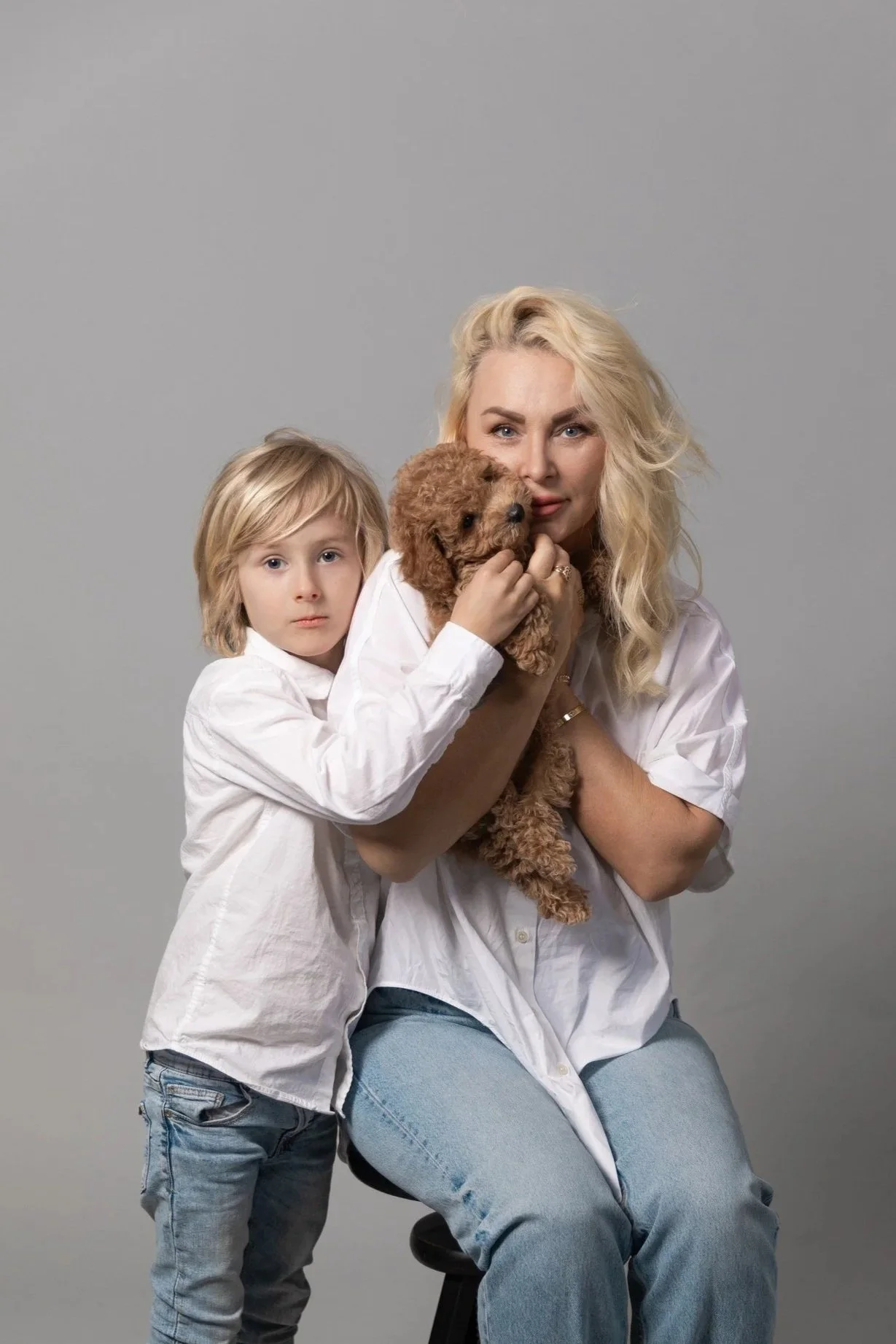 A woman and a young boy posing with a brown puppy against a plain gray background. The woman is sitting on a stool, holding the puppy close to her face, while the boy is leaning on her with his arm around her shoulder.