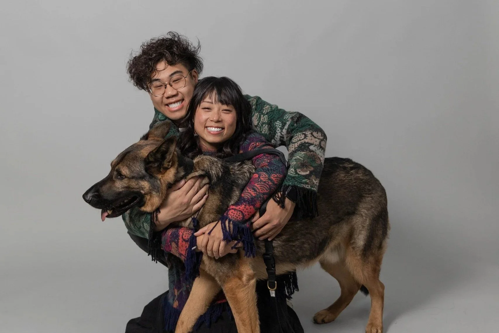 Two smiling people hugging a German Shepherd dog against a plain background.