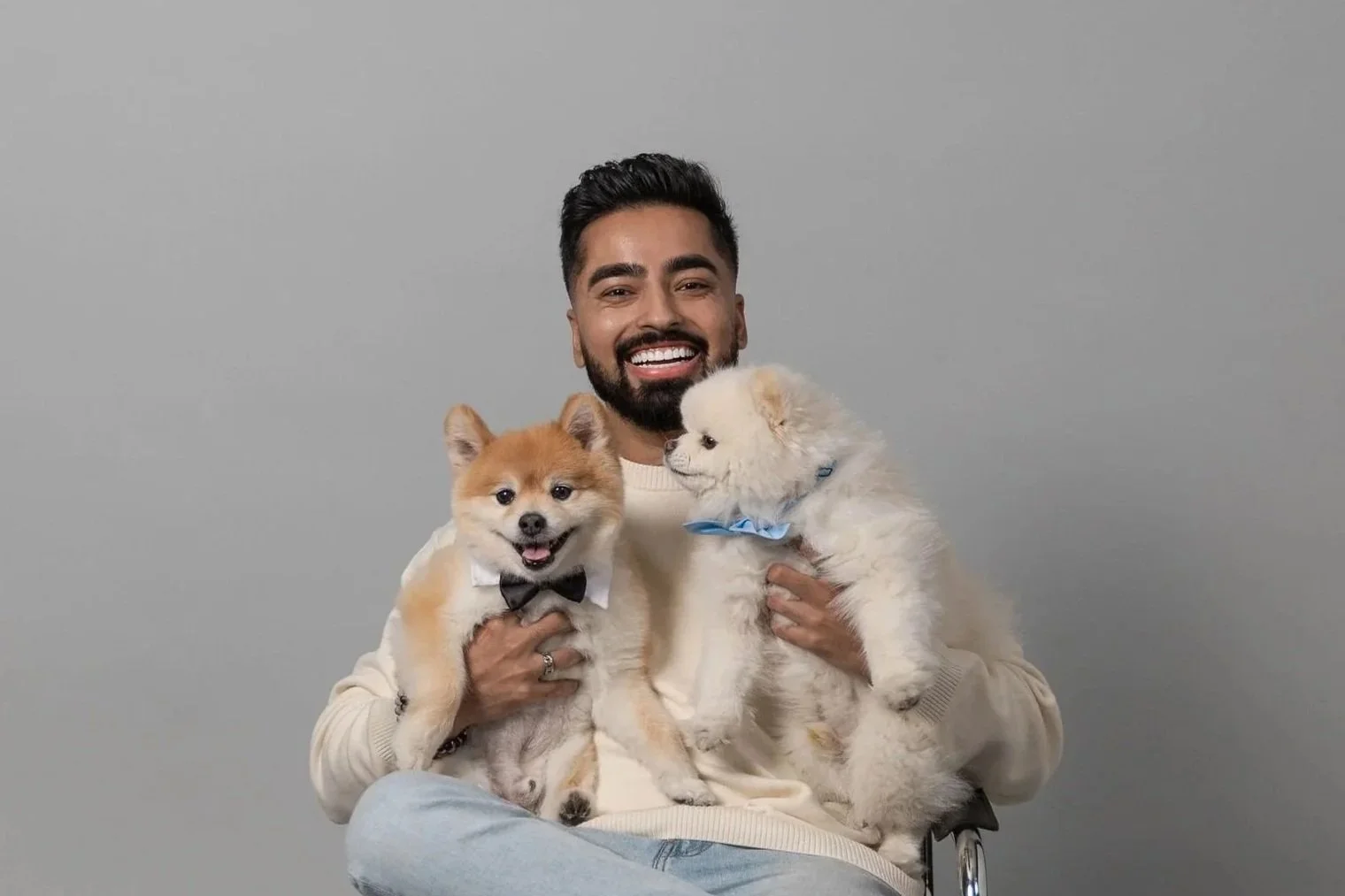 Smiling man holding two small dogs, one with a bow tie and the other with a blue bandana, against a plain gray background.