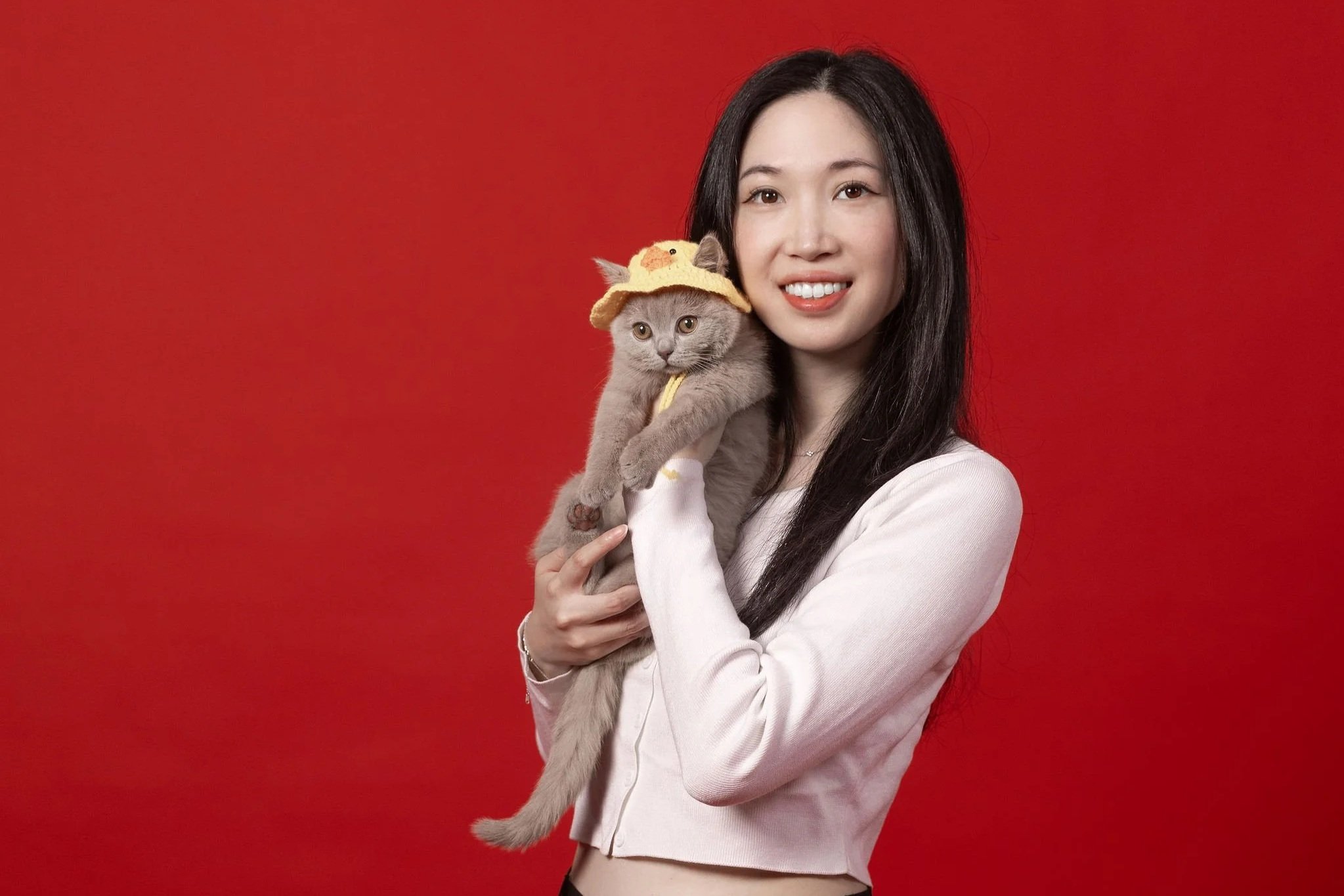 A woman holding a gray cat wearing a yellow chick hat against a red background.