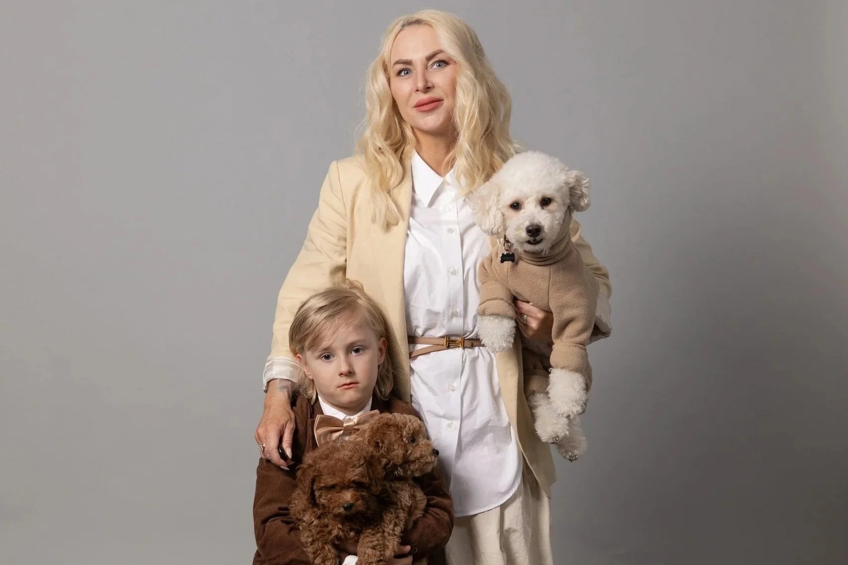 A woman with blonde hair and a beige blazer standing with two children, a girl and a boy, holding small poodle dogs against a plain grey background.