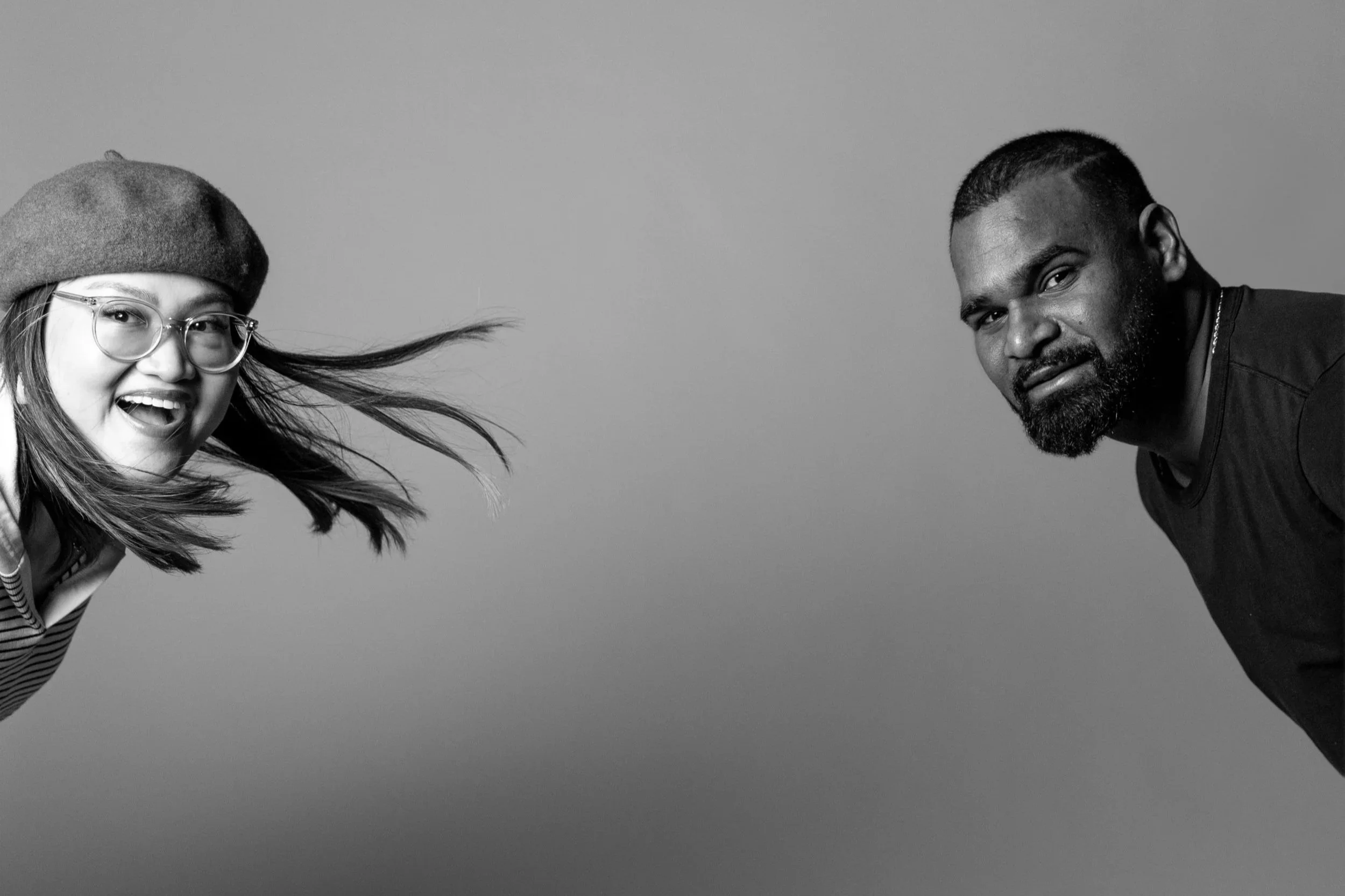 A black and white photo of a woman with glasses and a beret smiling and leaning towards the camera, and a man with a beard leaning in from the right side, both looking at the camera.