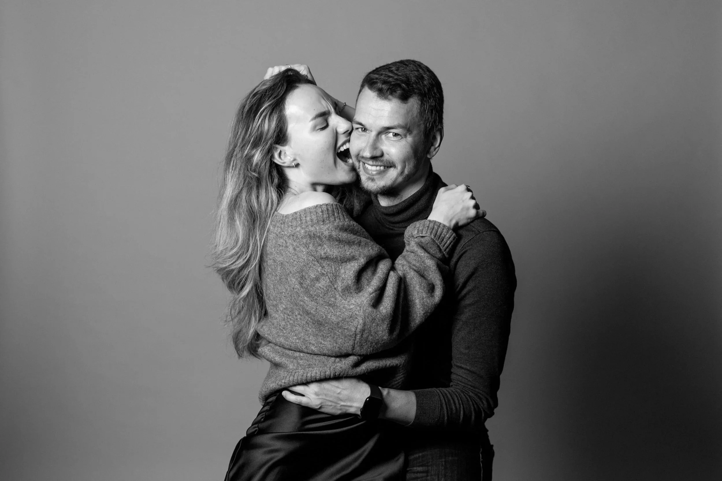 A black and white photo of a smiling man and woman in a close embrace. The woman is whispering into the man's ear, and the man is smiling at the camera. The woman has long wavy hair, and the man has short hair and a beard. They are wearing sweaters, 