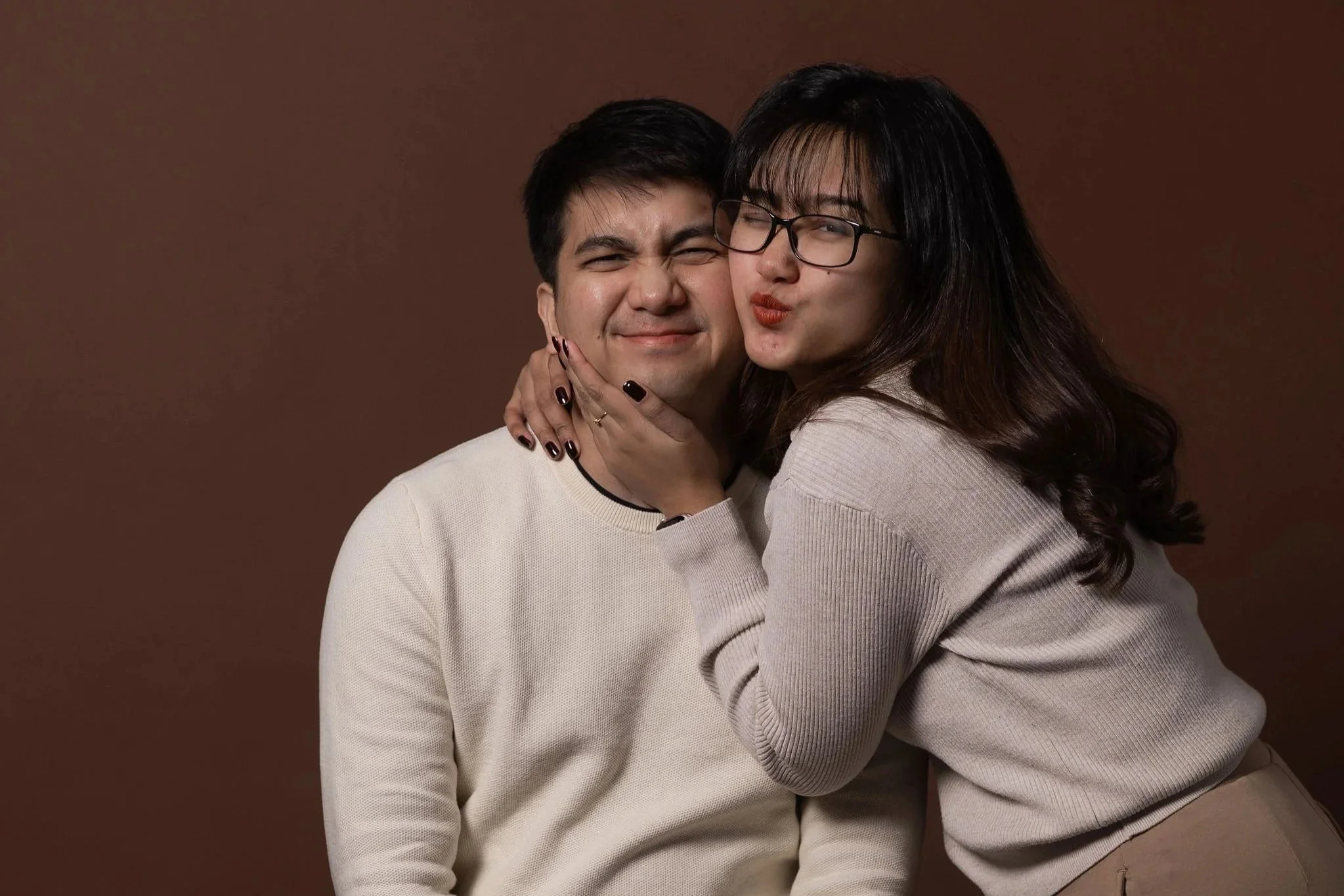A man and a woman posing together against a brown background. The man is smiling with his eyes closed, and the woman is making a pouting face while holding his face gently.