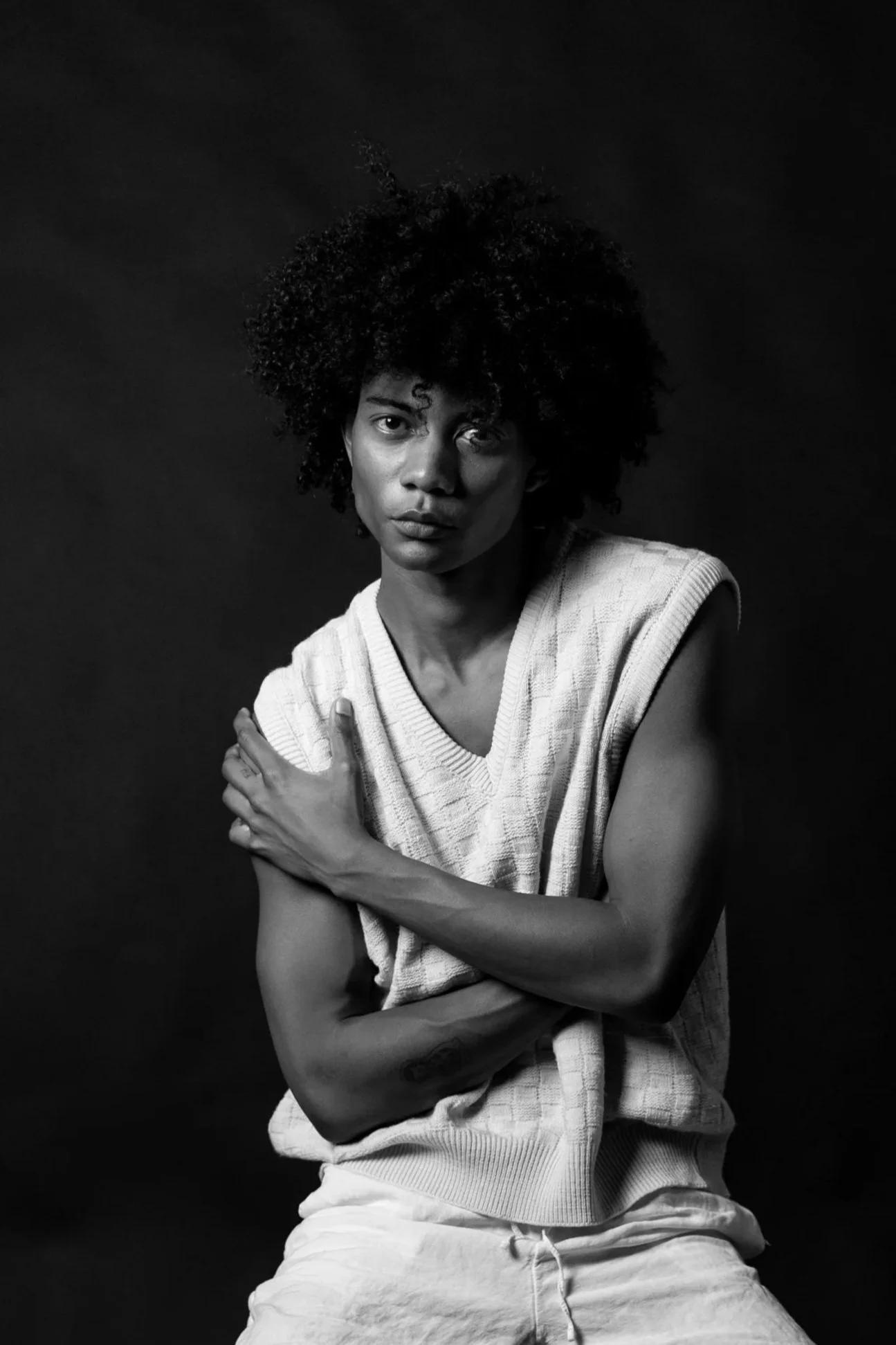 Black and white portrait of a woman with natural curly hair, wearing a sleeveless sweater and casual pants, looking directly at the camera against a plain dark background.