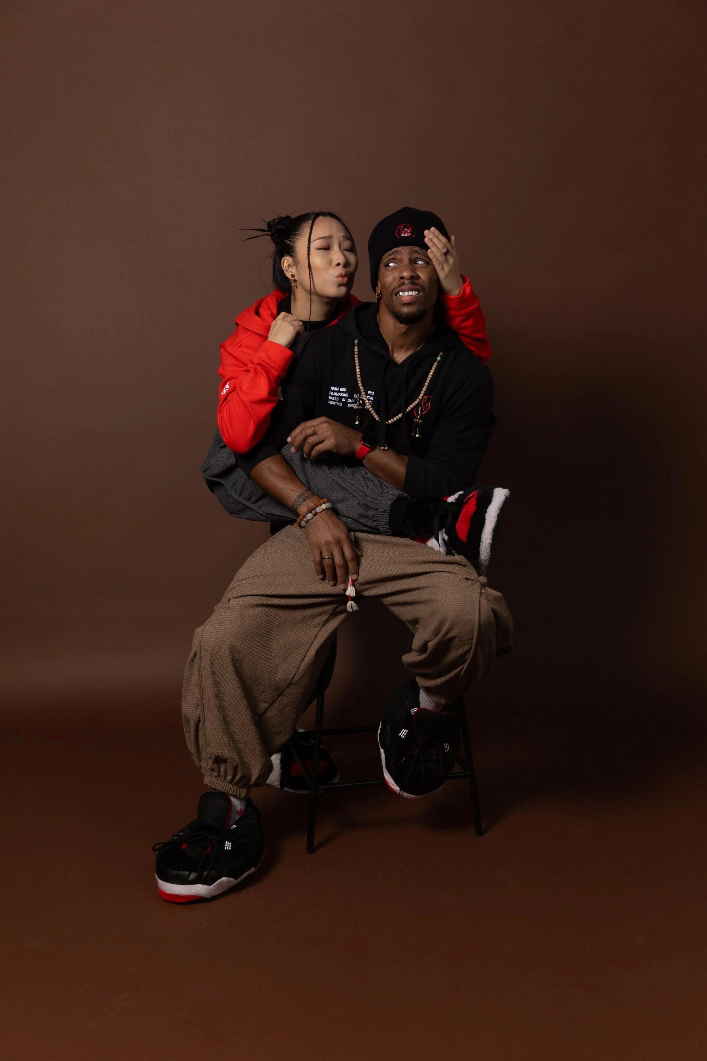 A woman with braids and earrings playfully holds a man's head while sitting on a stool against a brown background. The man, wearing a black hoodie, brown baggy pants, and sneakers, appears amused and is sitting with his hands crossed.