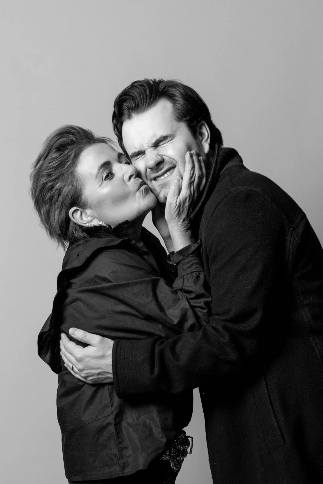 Black and white photo of a woman kissing a man on the cheek, both with their eyes closed, while the woman gently touches the man's face and the man has his eyes closed, showing an emotional moment.