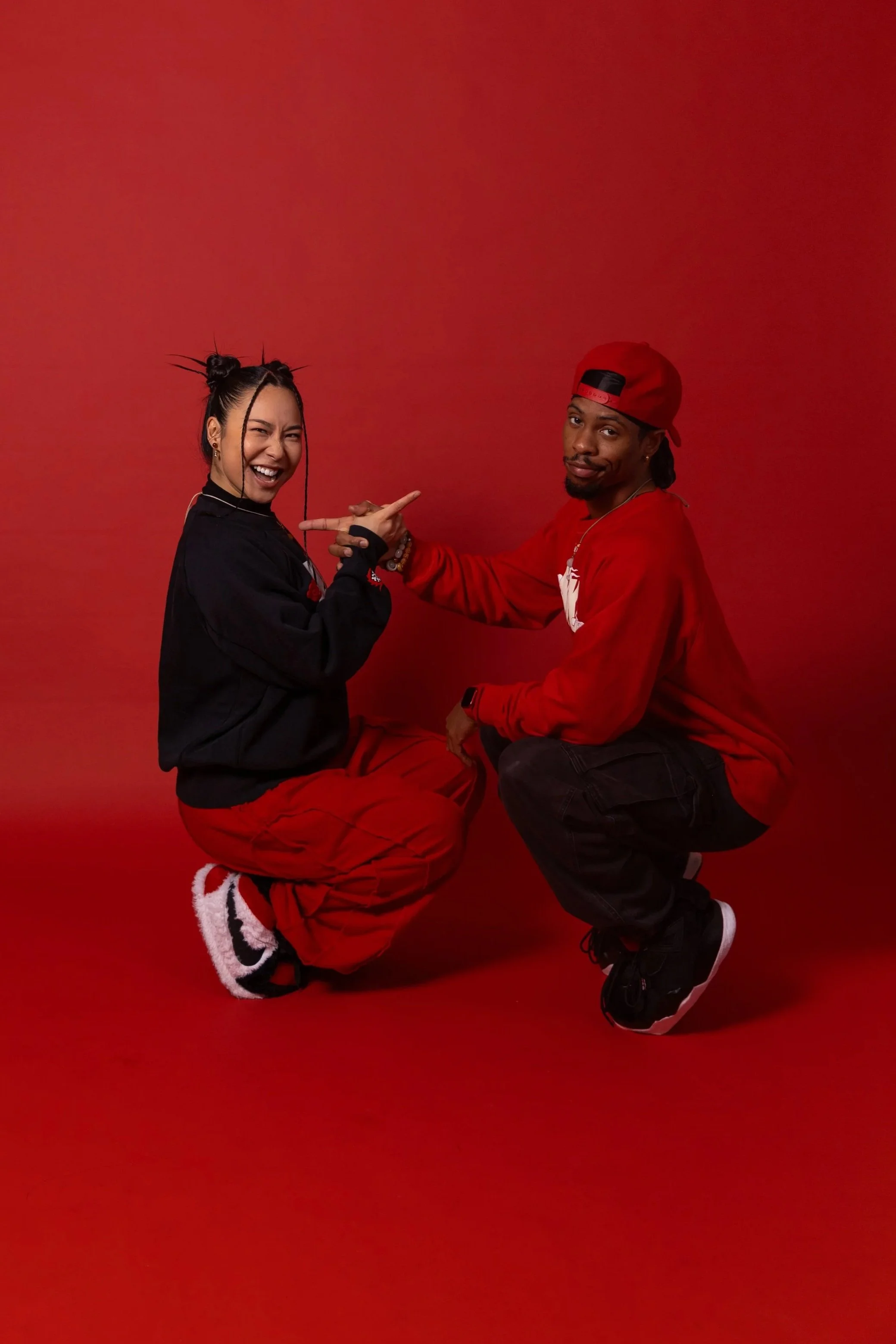 Two people kneeling on a red background, pointing at each other, smiling and making playful gestures. The woman on the left is wearing a black hoodie and red pants, and the man on the right is wearing a red sweatshirt, black pants, and a red cap.
