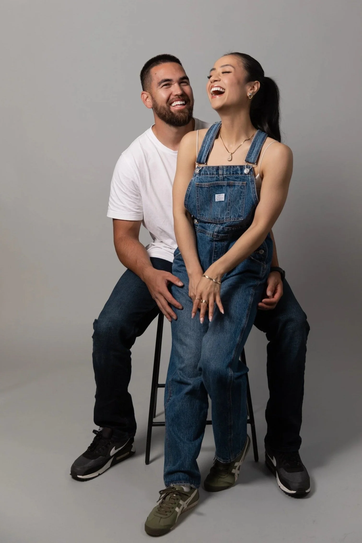 A man and woman sitting together, laughing and smiling, against a plain gray background.