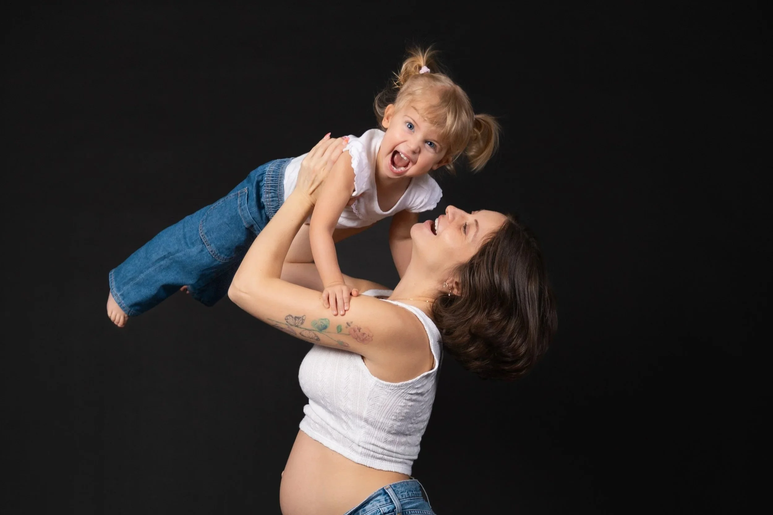 A woman with tattooed arms happily lifts a smiling young girl with pigtails in the air against a black background.