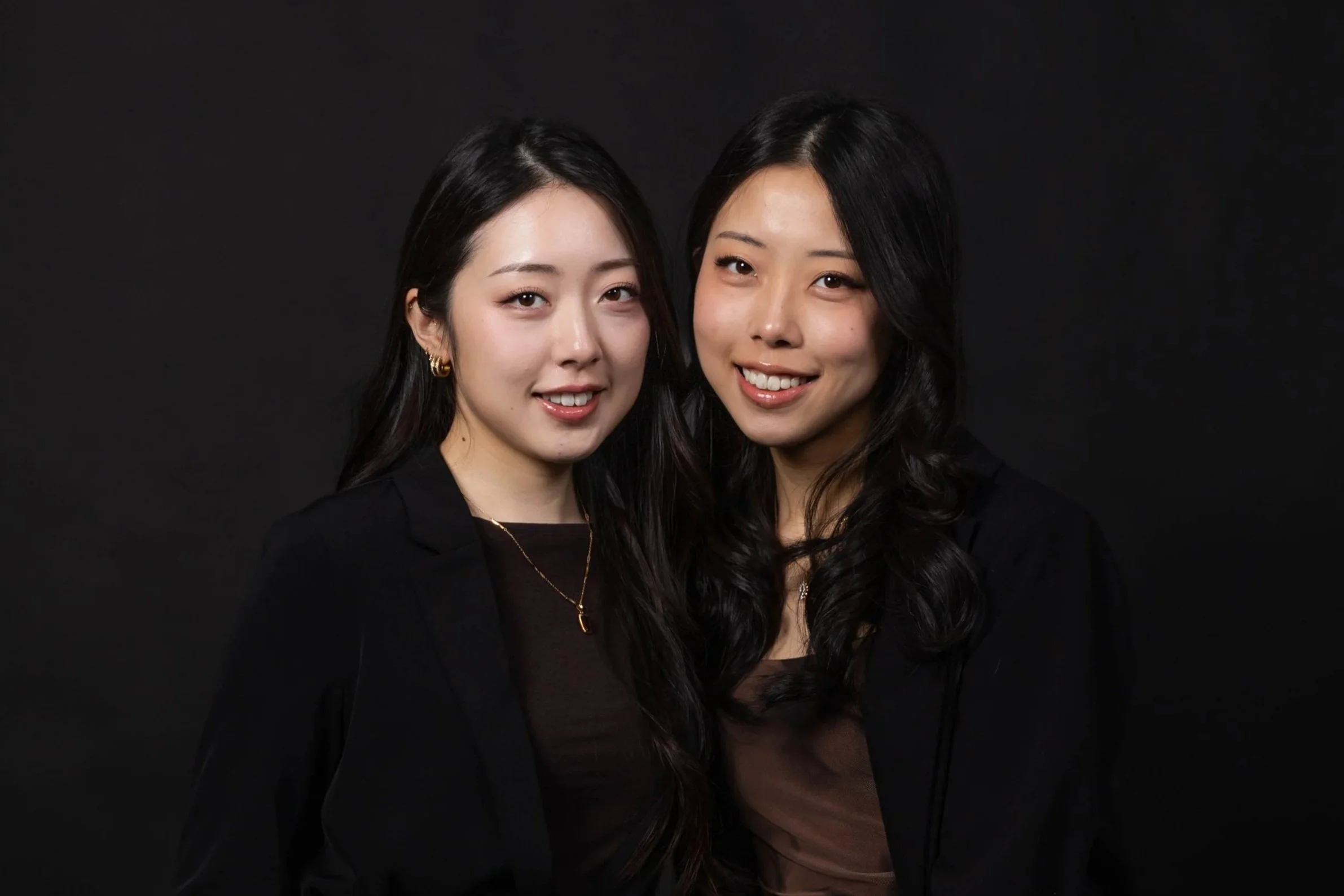 Two young women with dark hair, smiling and posing against a black background at a formal event.