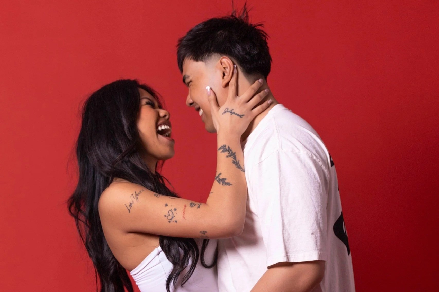 A happy couple with tattoos on their arms and shoulders, smiling and looking into each other's eyes, against a red background.