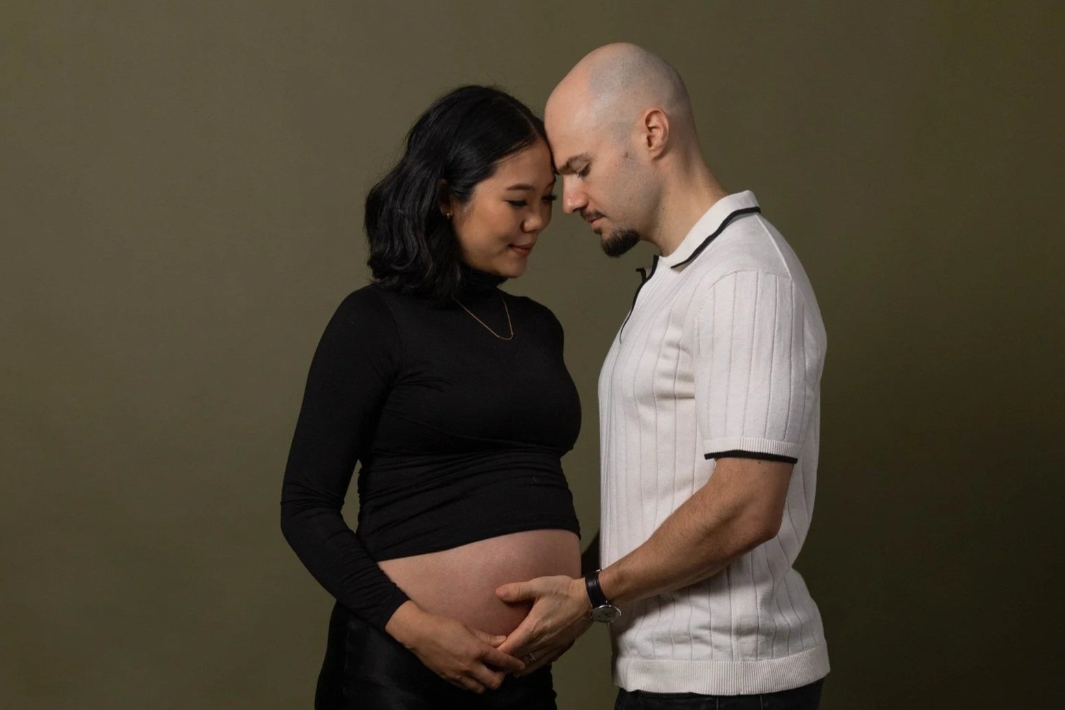 A pregnant woman with black hair and a man with a shaved head and a goatee standing close, touching her belly, both with their foreheads touching and eyes closed in a tender moment.