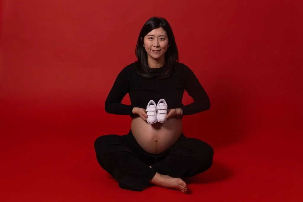 Pregnant woman sitting cross-legged against a red background, holding a pair of baby shoes in front of her belly.
