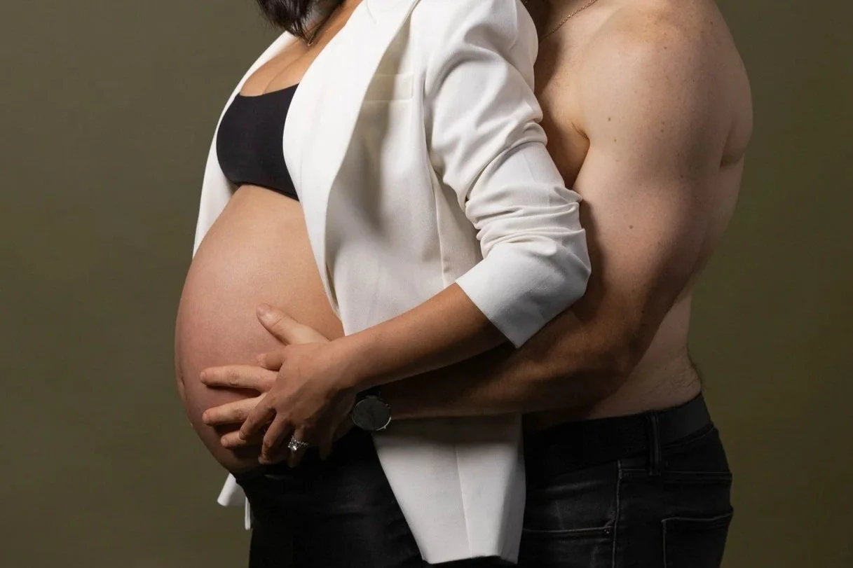 A pregnant woman with long dark hair wearing a black bra and white blazer, holding her belly with her right hand, standing in front of a shirtless man with muscular arms, visible chest, and a watch, against a plain background.