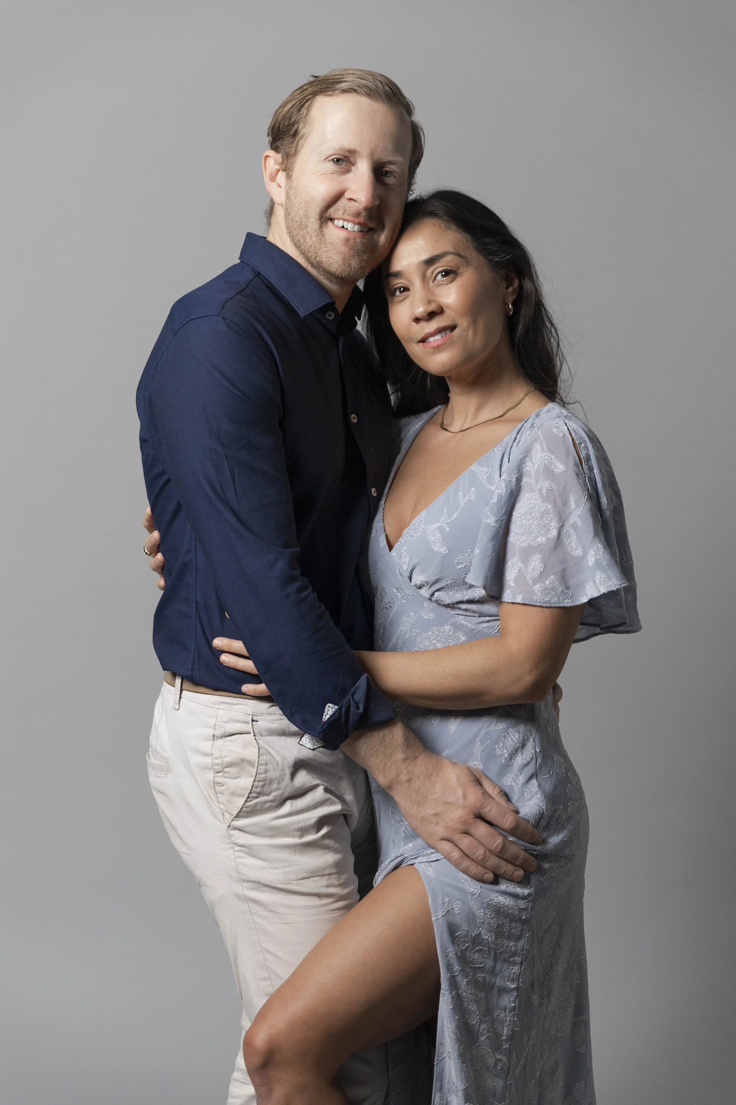 A couple standing close together: a man with light skin, wearing a navy blue shirt and beige pants, smiling, and a woman with medium skin, wearing a light blue floral dress, looking at the camera, with the man's arm around her waist and her leg sligh