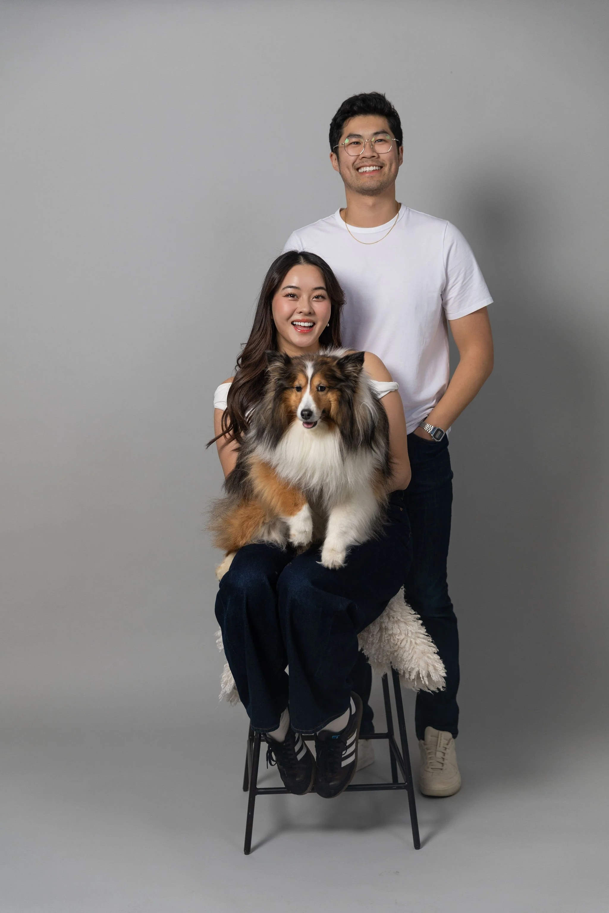 Two smiling young adults, one sitting on a stool with a Shetland Sheepdog, and the other standing behind them, all dressed casually against a plain gray background.