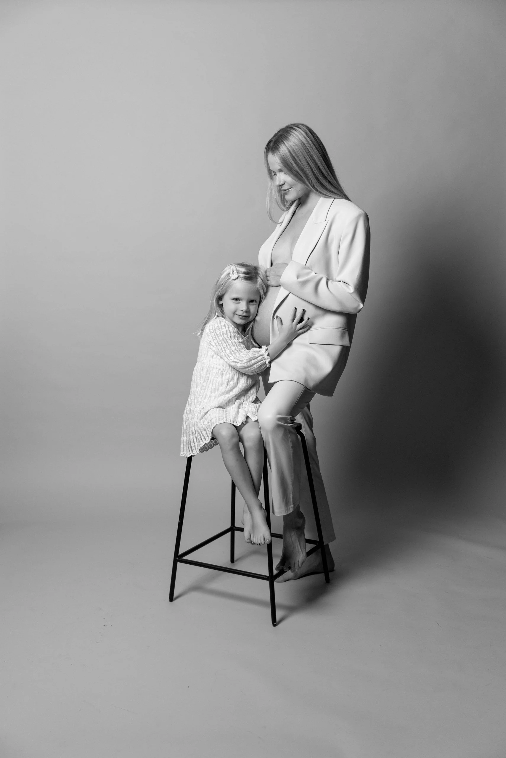 Black and white photo of a pregnant woman wearing a blazer and dress, sitting on a stool, with a young girl sitting on her lap, smiling and touching her belly.