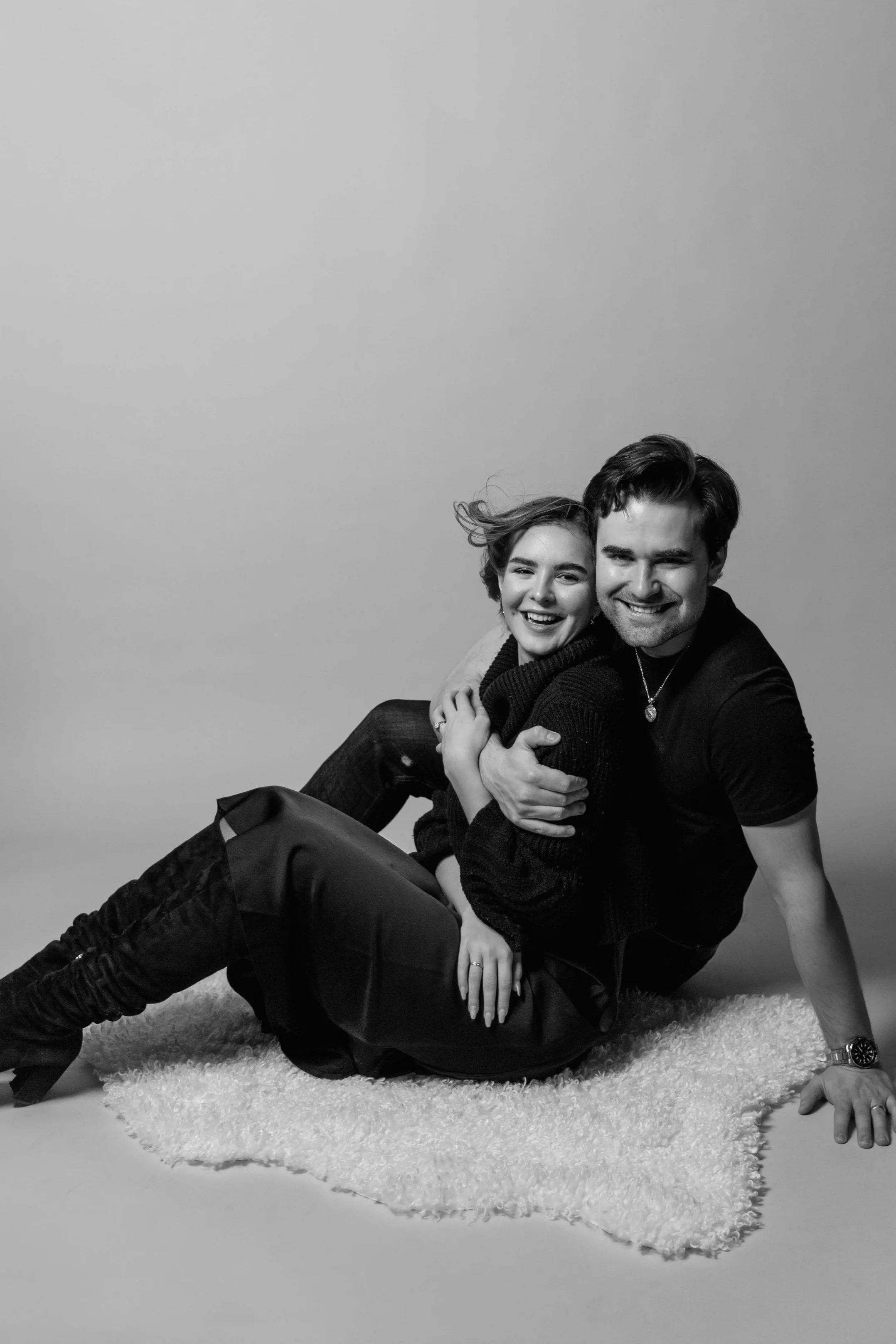 A black-and-white photo of a smiling young woman and man sitting on a fluffy white rug, hugging each other, with a plain light gray background.