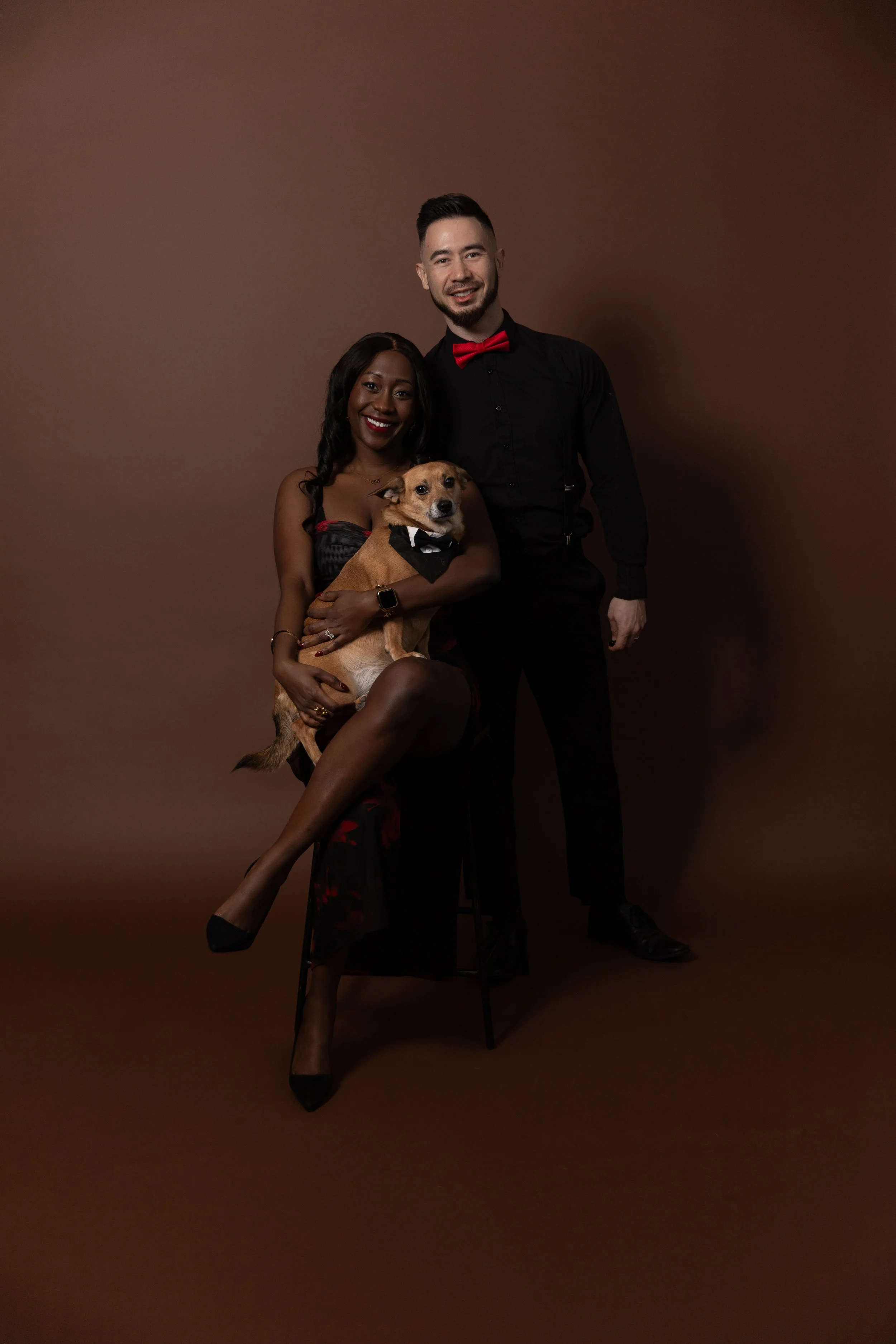 A man and woman dressed in formal attire with a dog sitting on the woman's lap against a brown background.