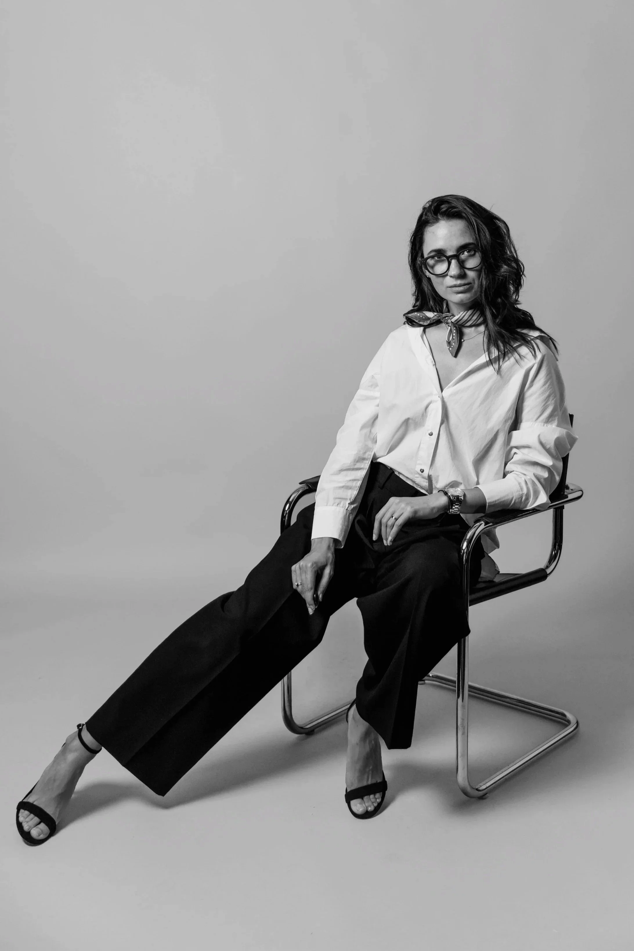 A woman sitting on a modern chair, dressed in a white blouse and black wide-leg pants, wearing glasses and a scarf around her neck, in a black and white studio shot.