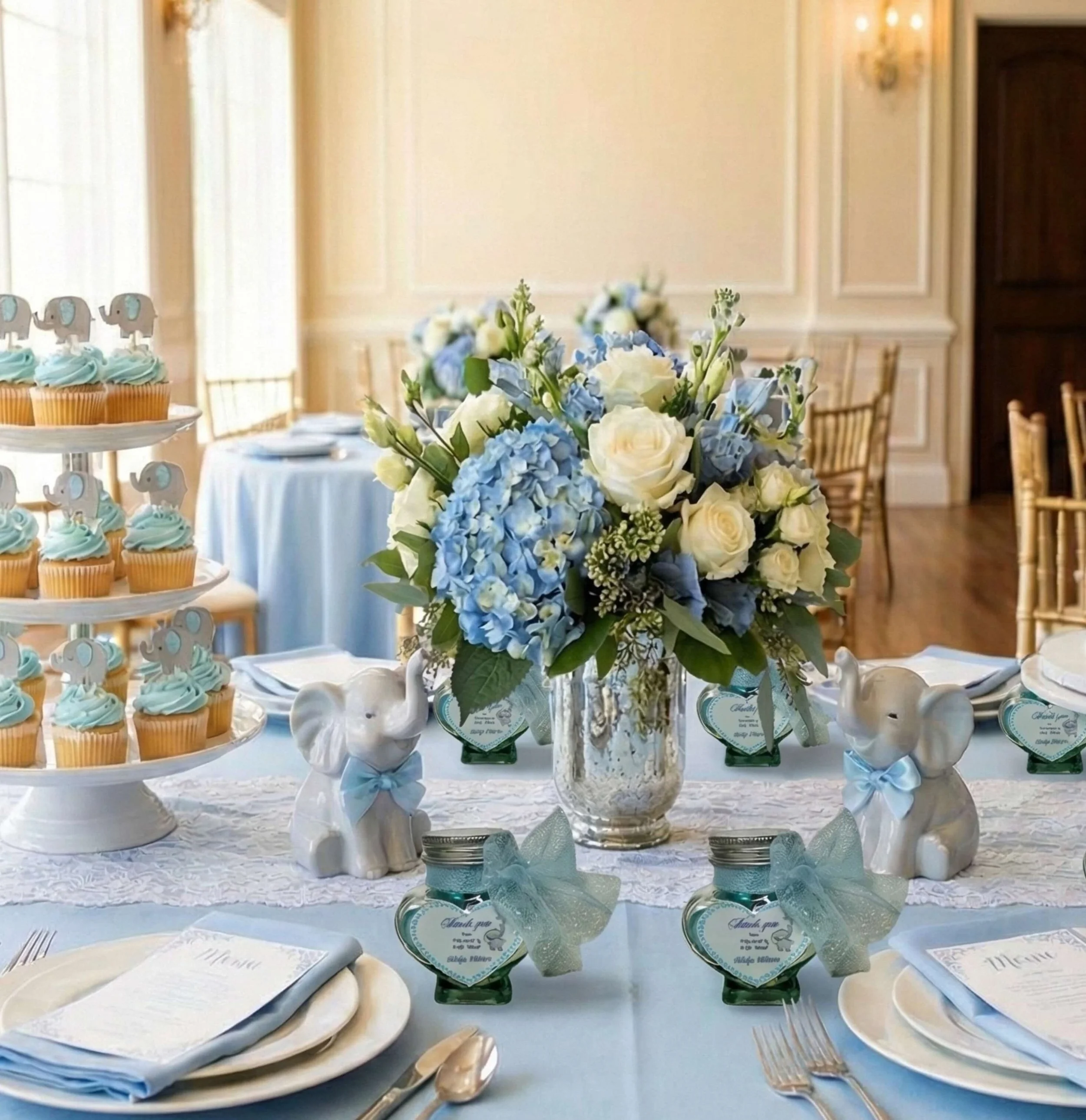 Table decorated with a blue and white theme for a celebration, featuring a floral centerpiece with white and blue flowers, ceramic elephant figurines with blue bows, small jars with thank you tags, cupcakes with elephant toppers, plates, napkins, and utensils.