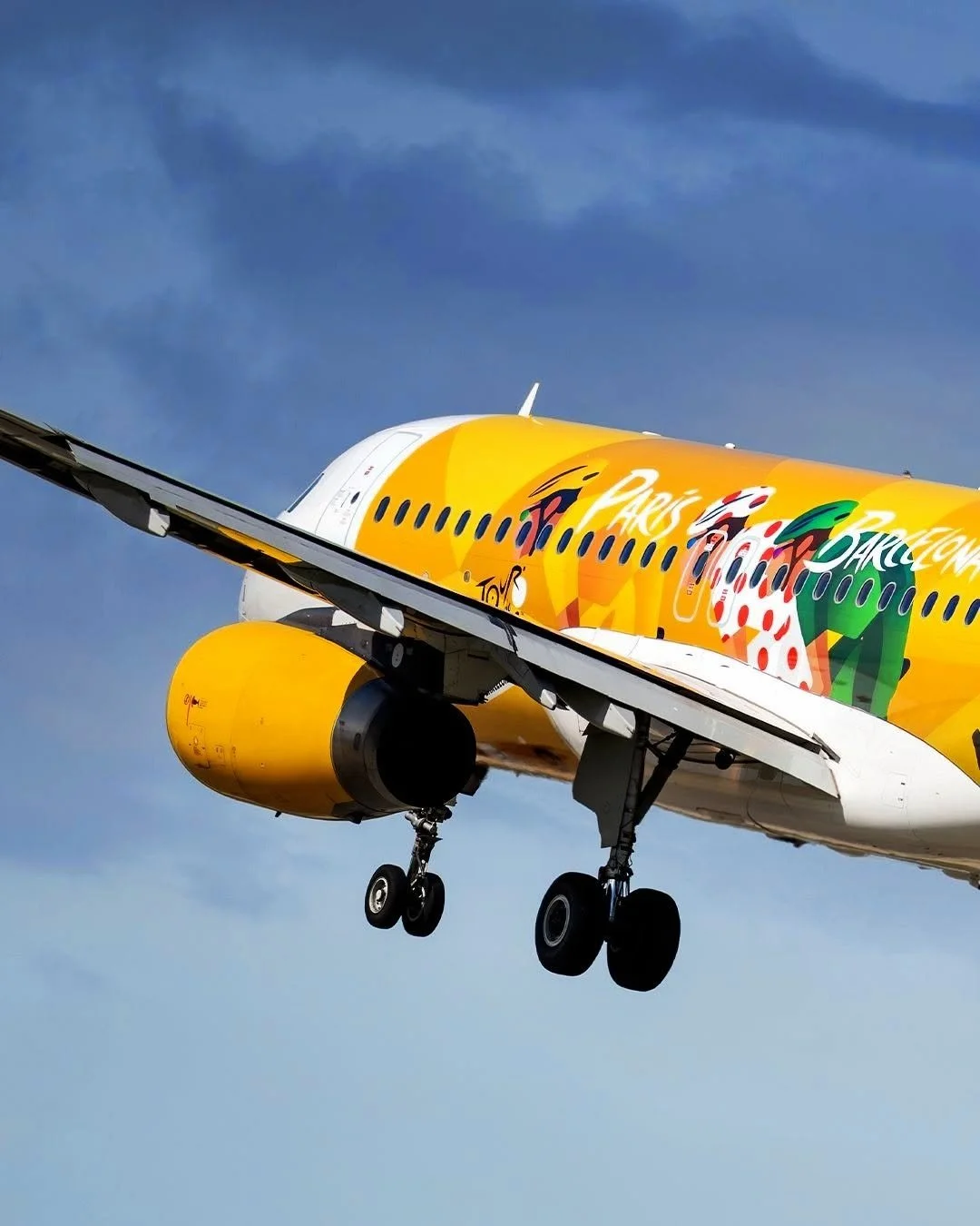 A colorful airplane with a yellow body and festive holiday graphics on the fuselage, flying in a partly cloudy sky.